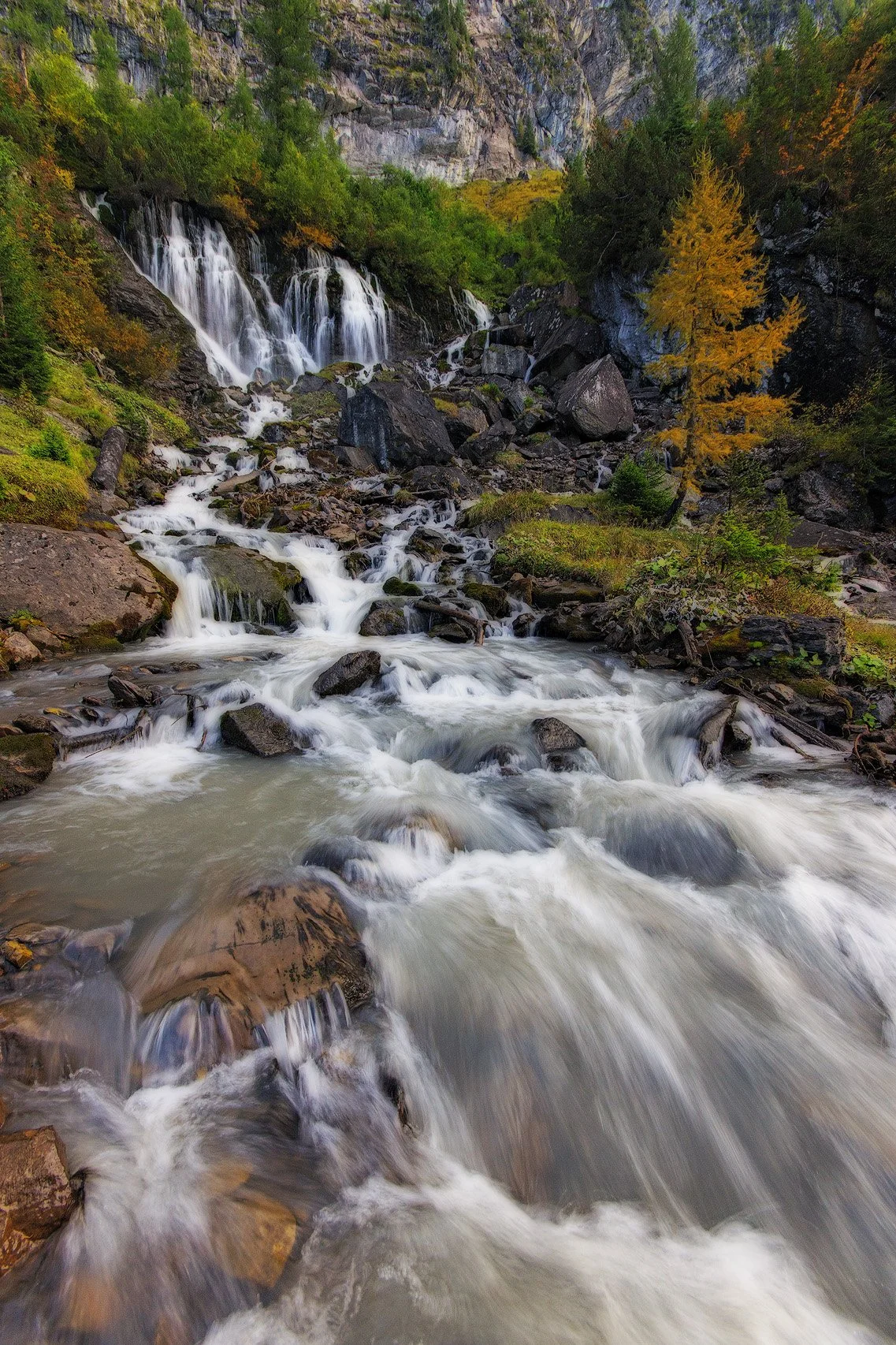 A cascading mountain stream flowing down a rocky slope with waterfalls, surrounded by trees with some showing fall foliage, and a steep rocky cliff in the background.