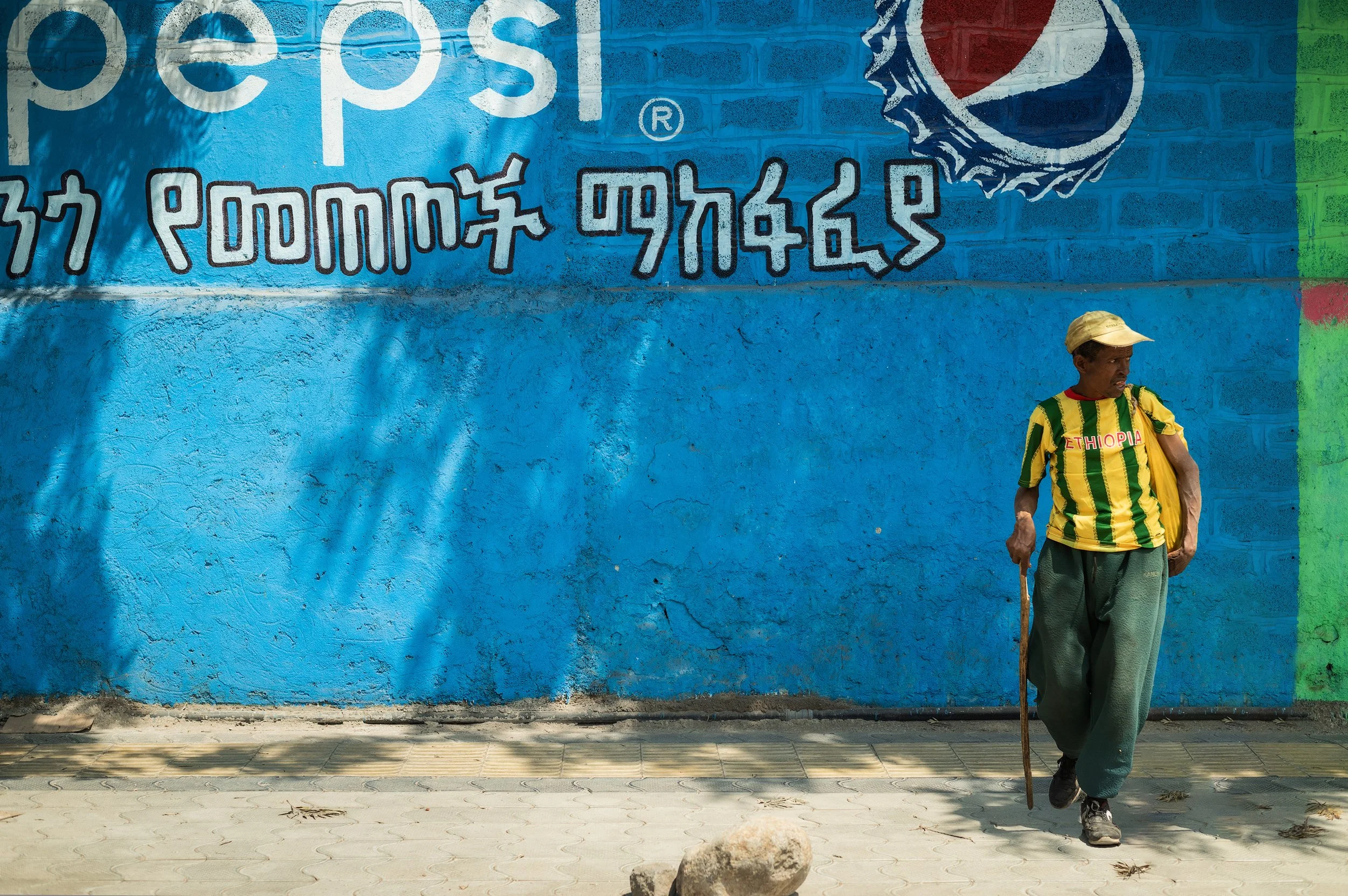 A man walking with a cane in front of a bright blue wall with graffiti, wearing a yellow and green striped Ethiopia jersey and a yellow cap.