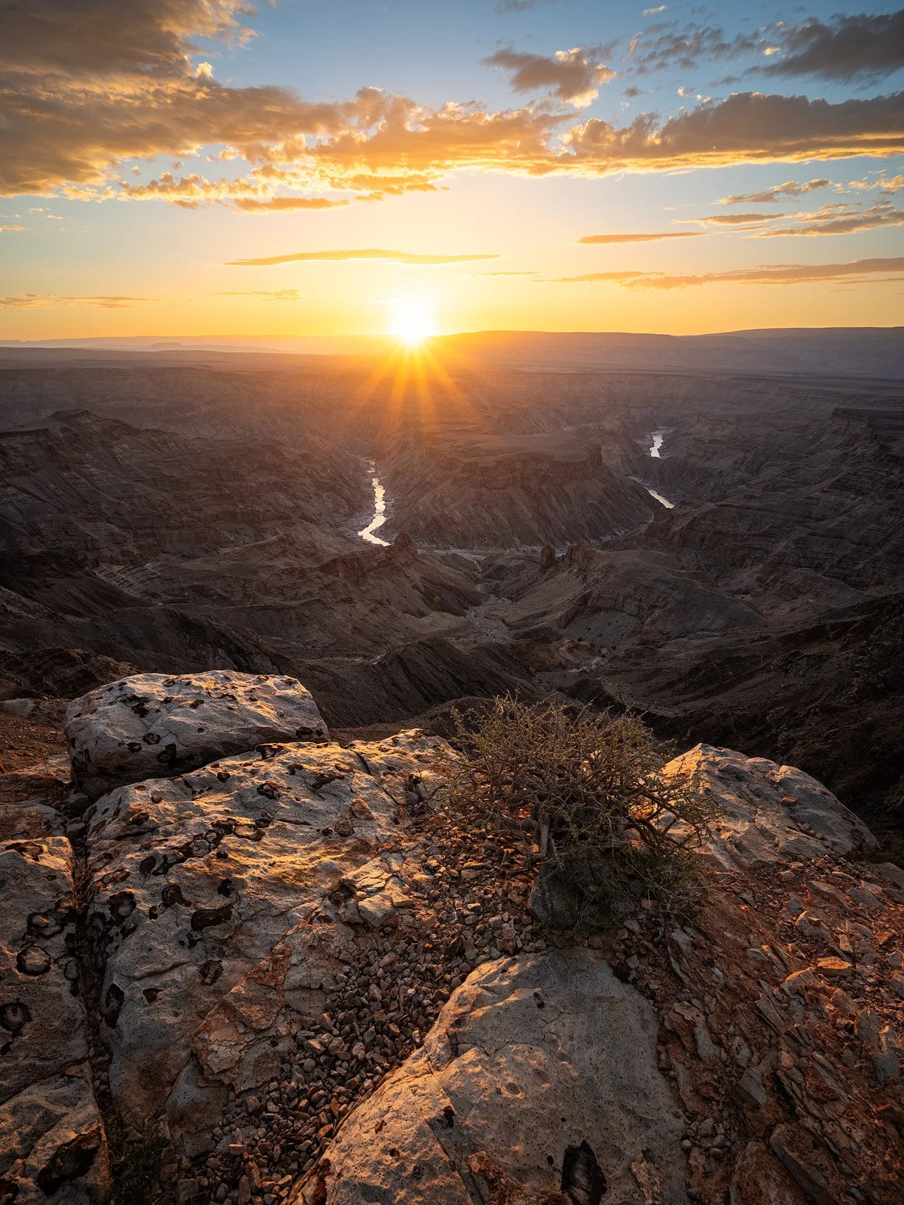 Sunset over the Grand Canyon with rocky foreground and cloudy sky.