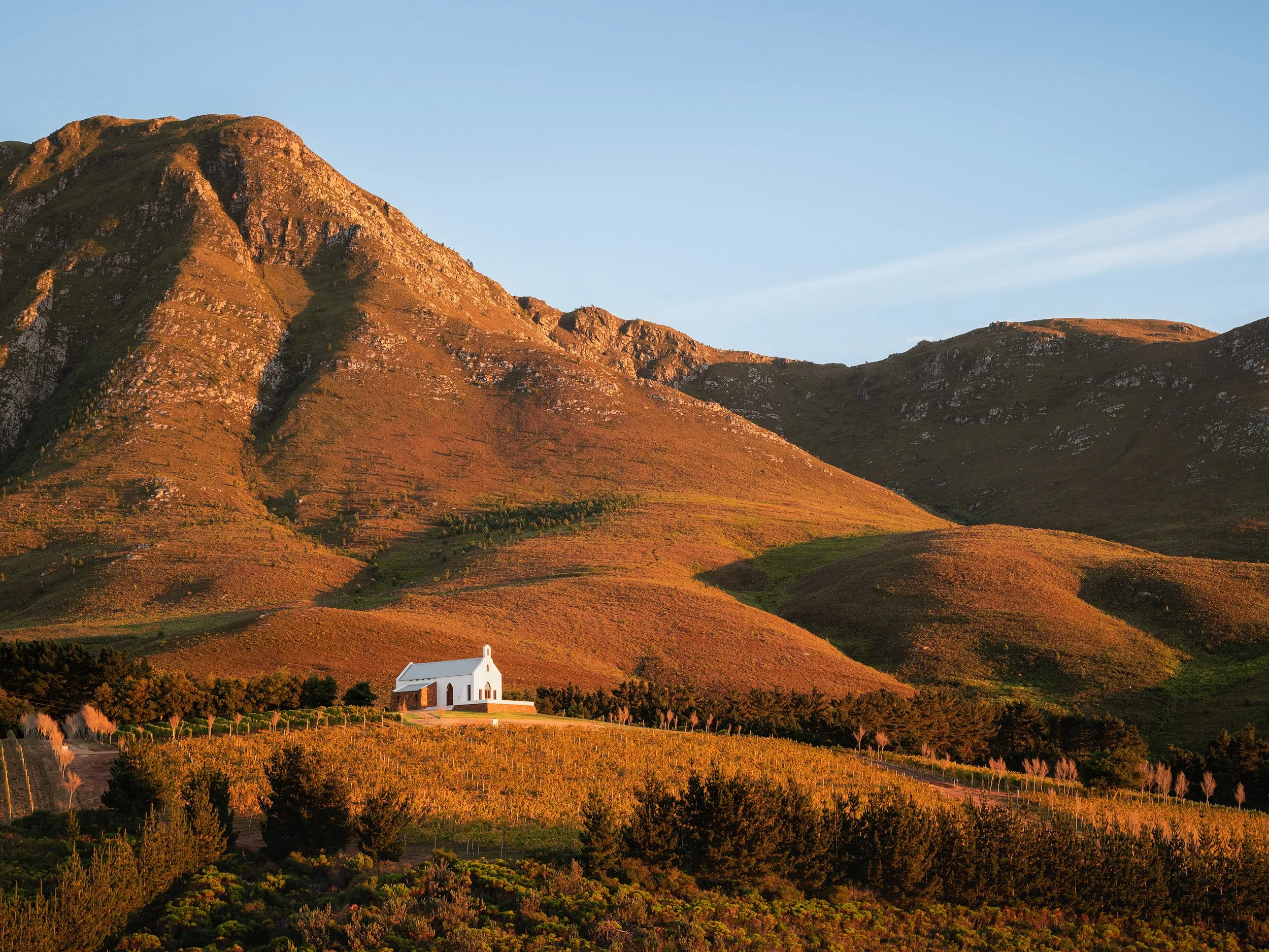 A small white church with a steeple located at the base of a large mountain range during sunset. South Africa, Hemel en Aarde, Winery, Ataraxia. 