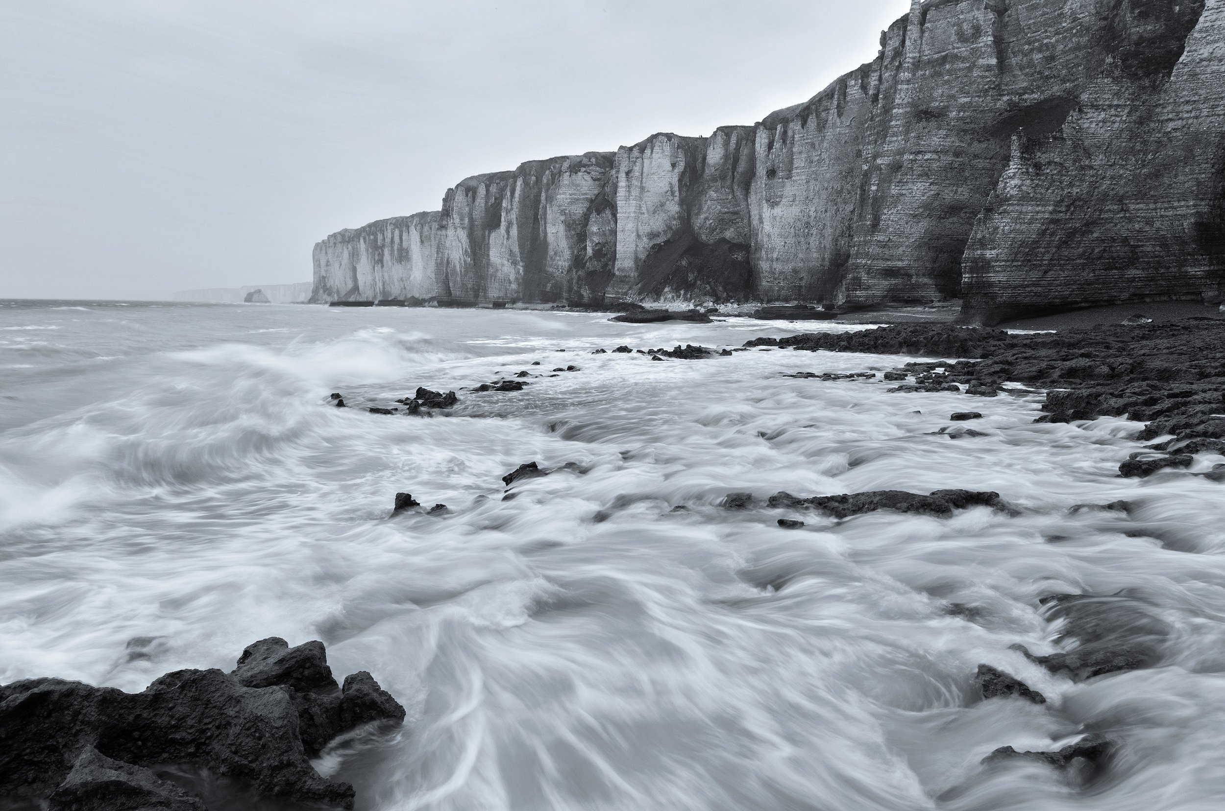 Black and white photograph of a rugged coastline with high chalk cliffs and waves crashing against rocks.