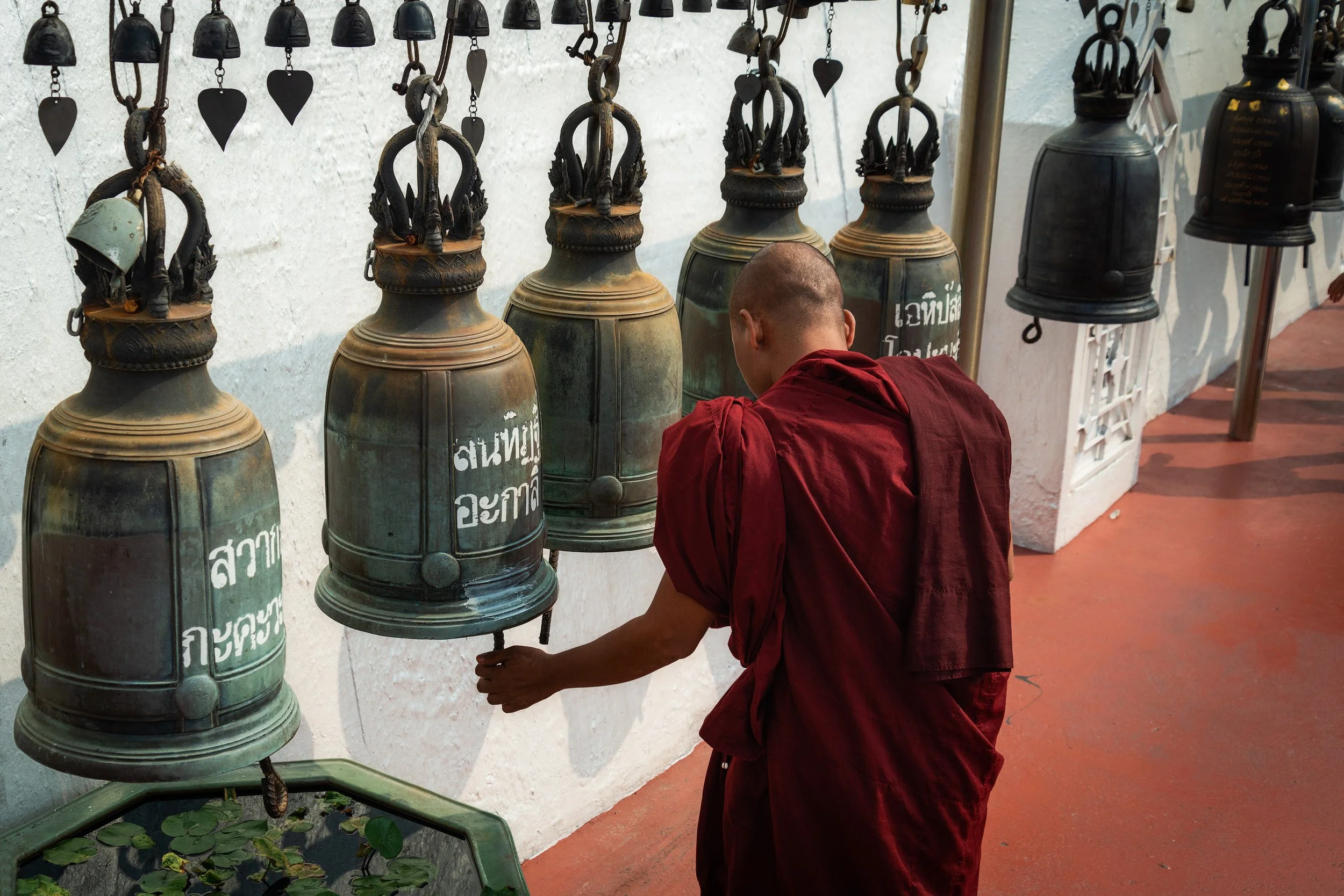 A young Buddhist monk dressed in red robes rings a large bronze bell hanging on a wall, with several other similar bells nearby in a temple setting.