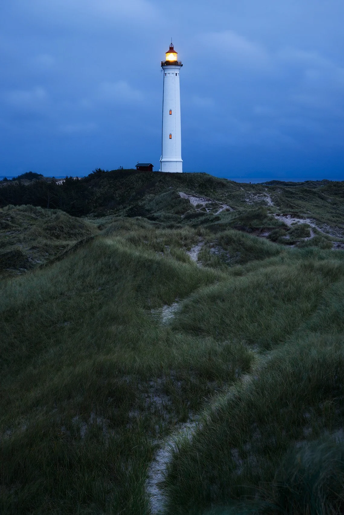 Lighthouse on a grassy hill during dusk with cloudy sky in the background.