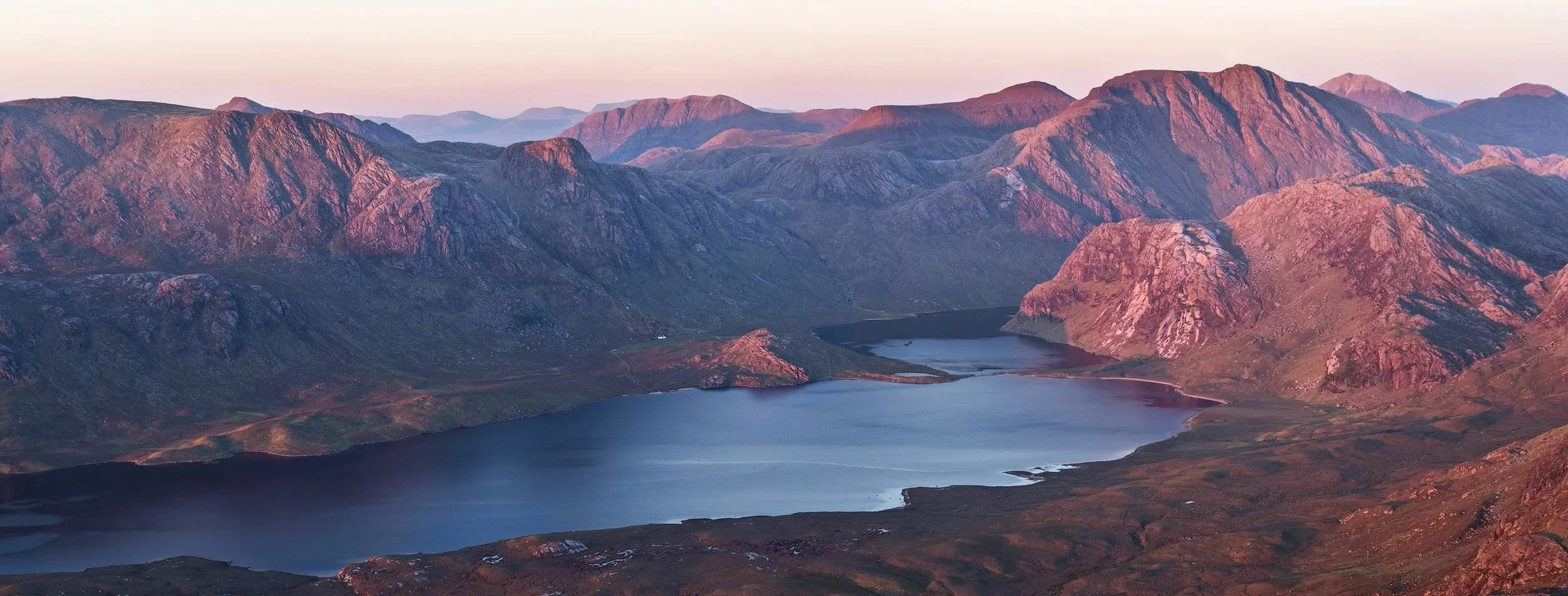 A panoramic view of Scottish mountains surrounding a lake at sunset, with pink and purple hues illuminating the peaks. Scotland. Highlands.