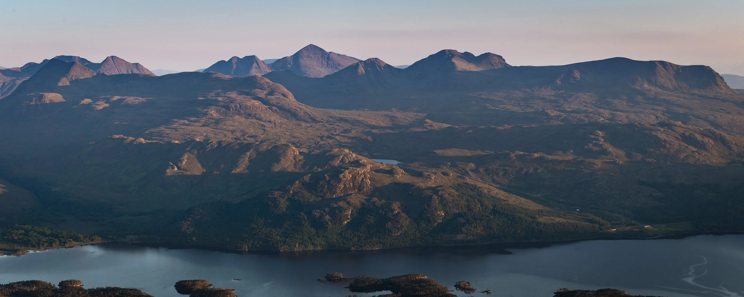 Mountain landscape with rolling hills, rugged peaks, and a body of water in the foreground.