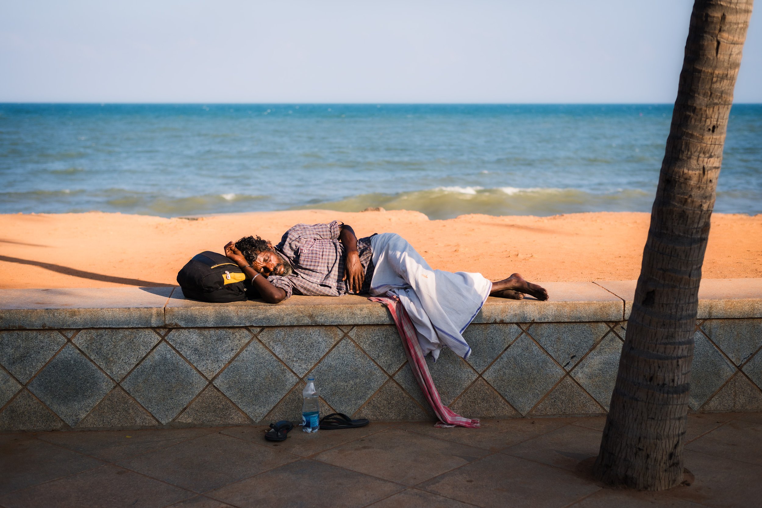A man with dark curly hair and a beard sleeping on a bench near the beach, with water and sand in the background. There is a black bag next to his head, and a plastic water bottle, flip-flops, and a piece of cloth on the ground nearby. A palm tree is