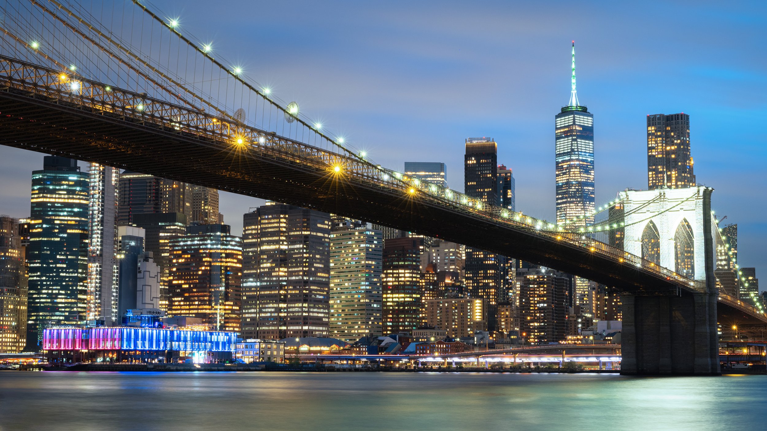 Night view of Brooklyn Bridge over Manhattan skyline with lit buildings including One World Trade Center.
