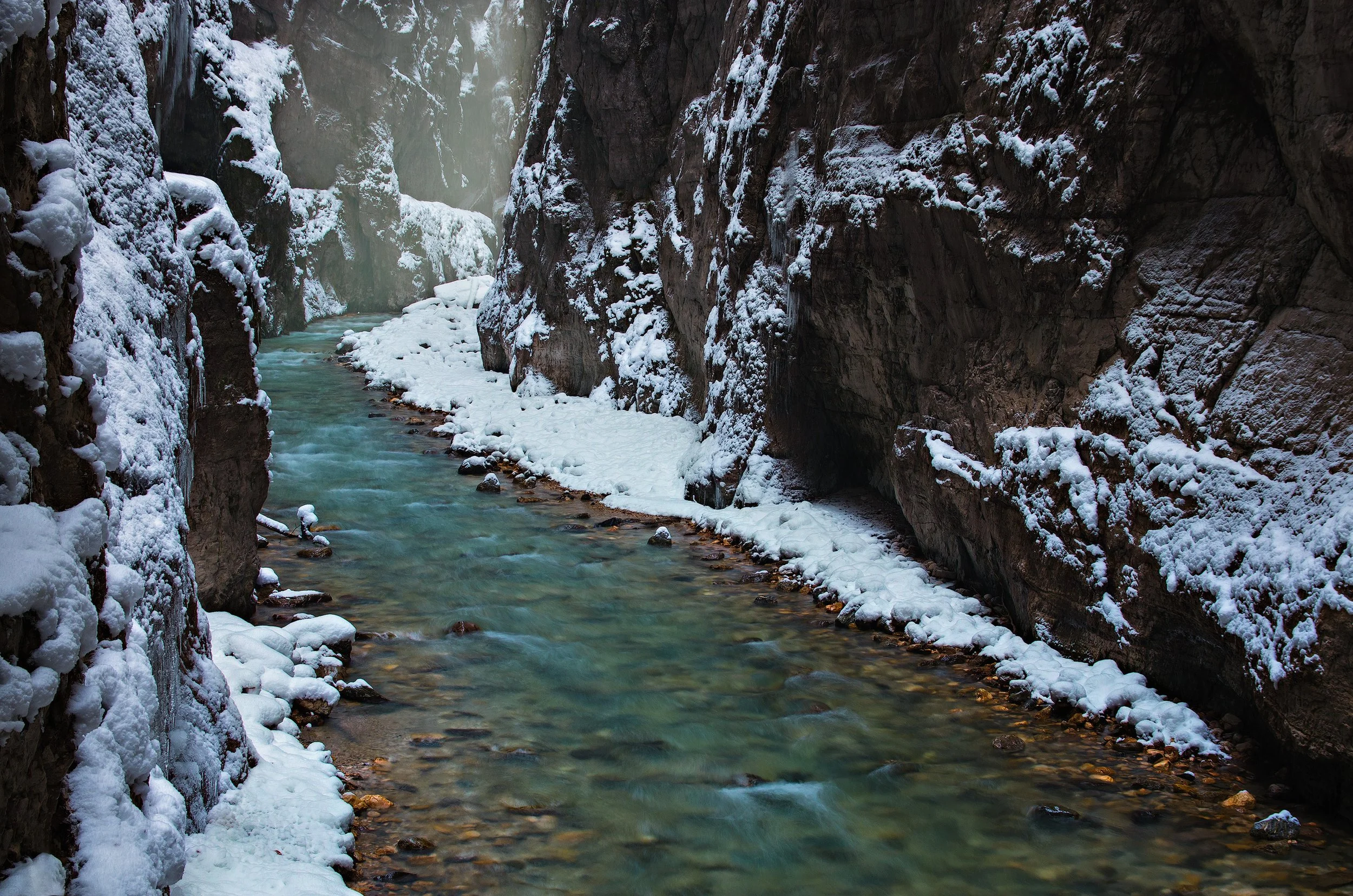 Narrow Partnachklamm in Bavaria with snow-covered rocks on the sides and a flowing turquoise river at the bottom, with mist and rocky walls.