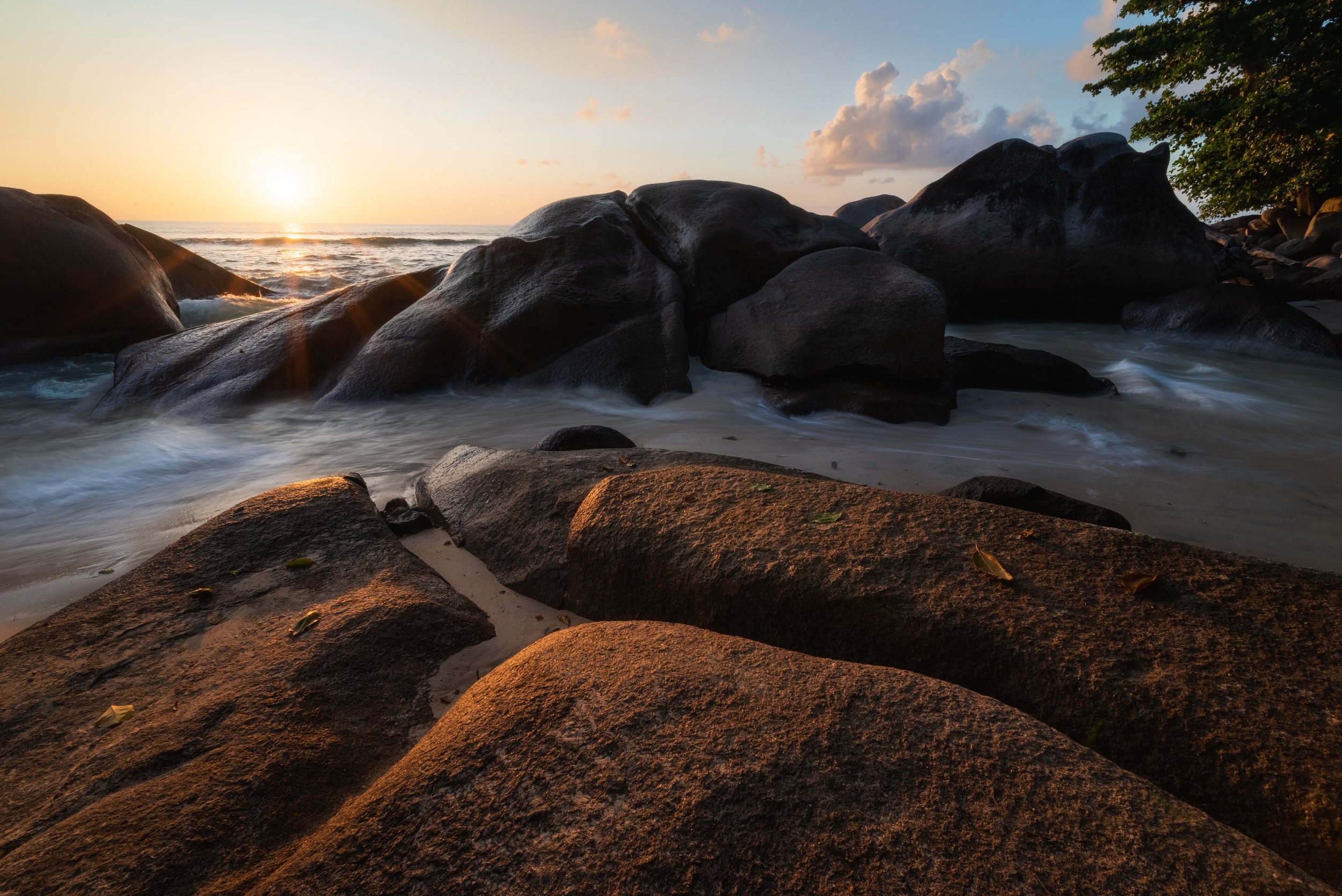 Sunset over a rocky beach with boulders, waves, and a tree on the right.