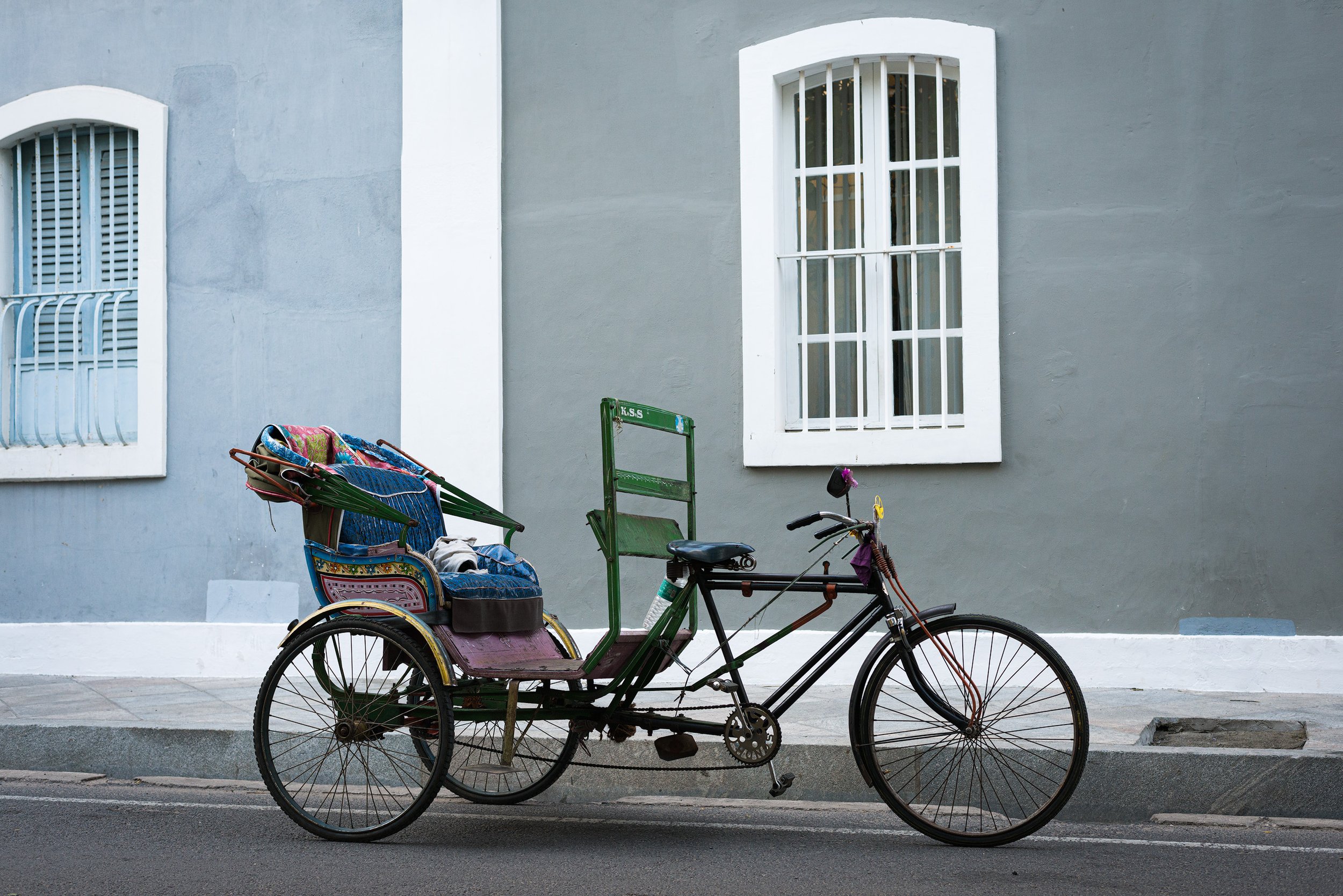 A three-wheeled bicycle rickshaw parked on the street in front of a gray building with two white-framed windows