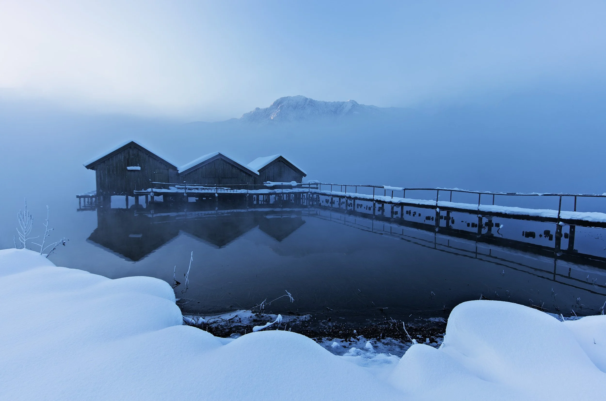 Snow-covered landscape with three wooden stilt houses reflecting in calm water, with a mountain in the background and overcast sky. Image was taken at Kochelsee in Bavaria.