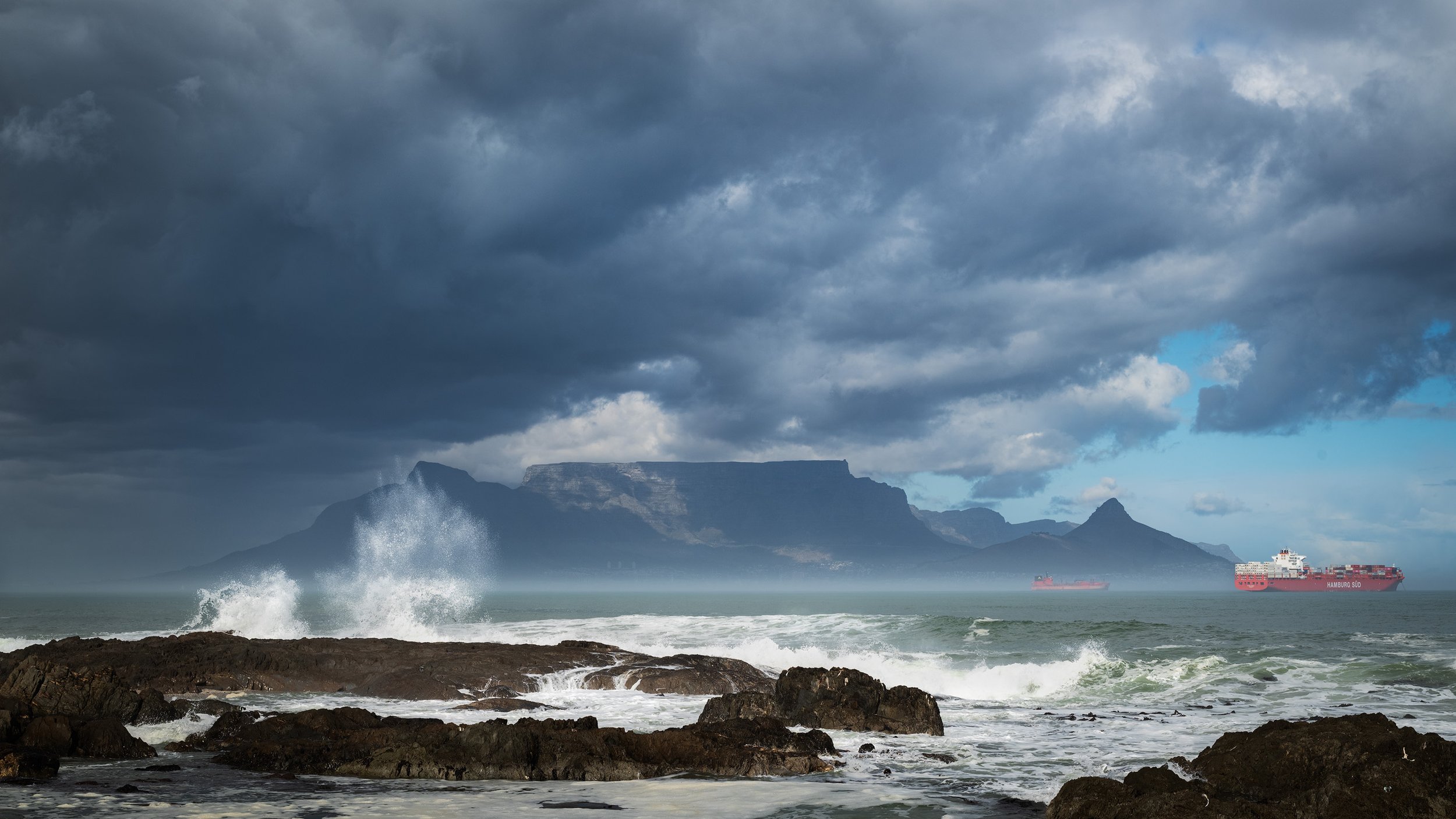 Stormy sky over a rocky coastline with waves crashing, mountains in the distance, and ships on the water.