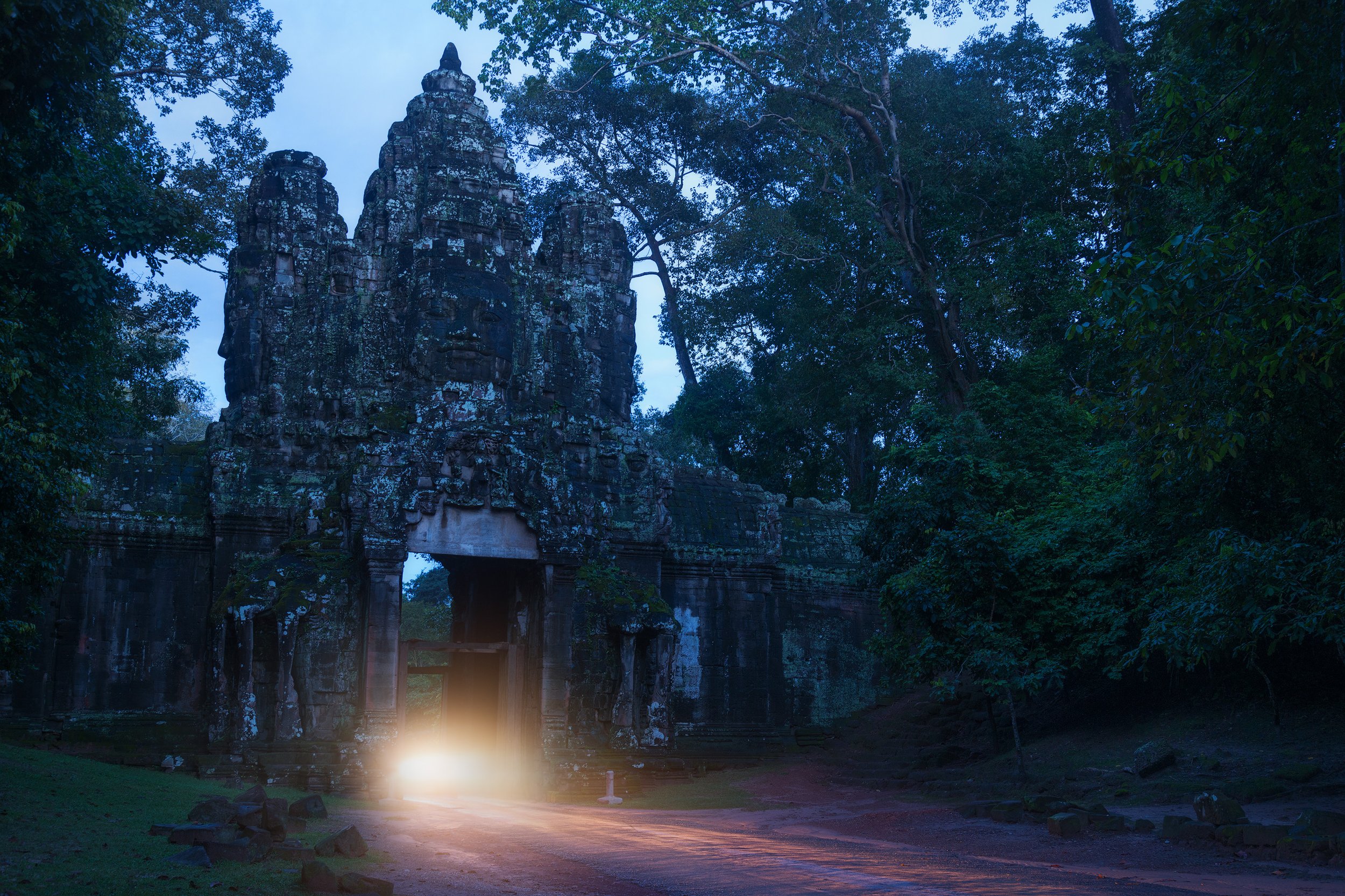 Ancient stone temple gate partially illuminated by a bright light in a forested area during dusk. Angkor Wat Cambodia.