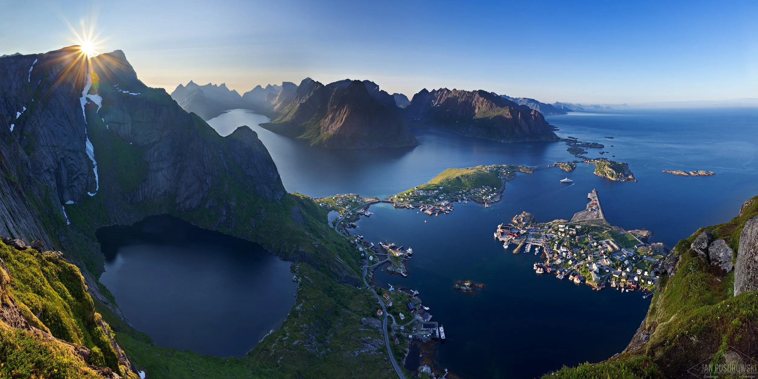 A scenic aerial view of a fjord landscape during sunset, featuring mountains, water, and a small town on the coast. Reinebringen, Reine, Norway, Lofoten.