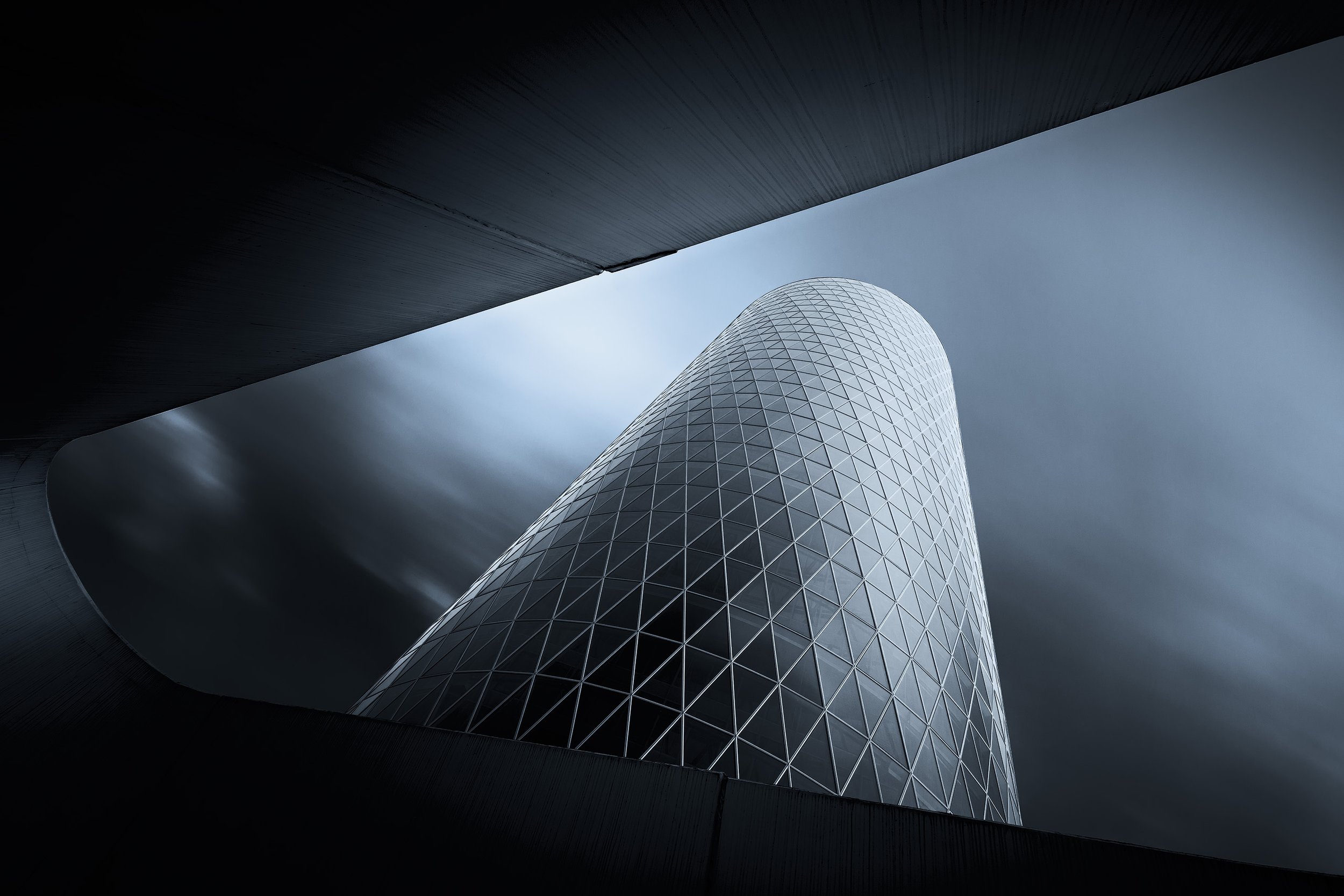 A modern skyscraper with a cylindrical shape and glass facade seen from below, framed by dark architectural elements with cloudy sky background.