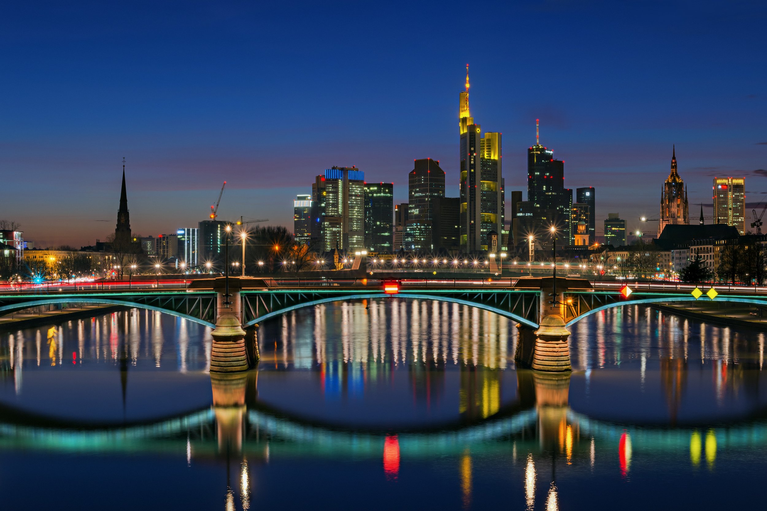 Night view of a city skyline with illuminated skyscrapers, bridges over a river, and reflections on the water.