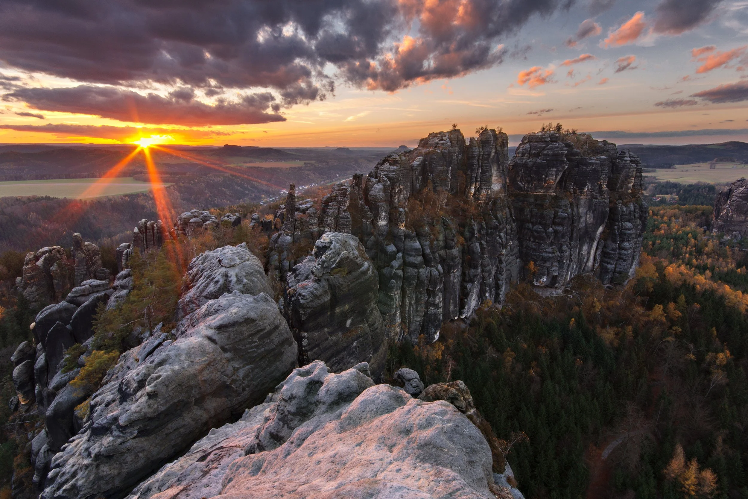 Sunset over rocky formations in a forested landscape, with clouds in the sky and trees with autumn foliage in the background.