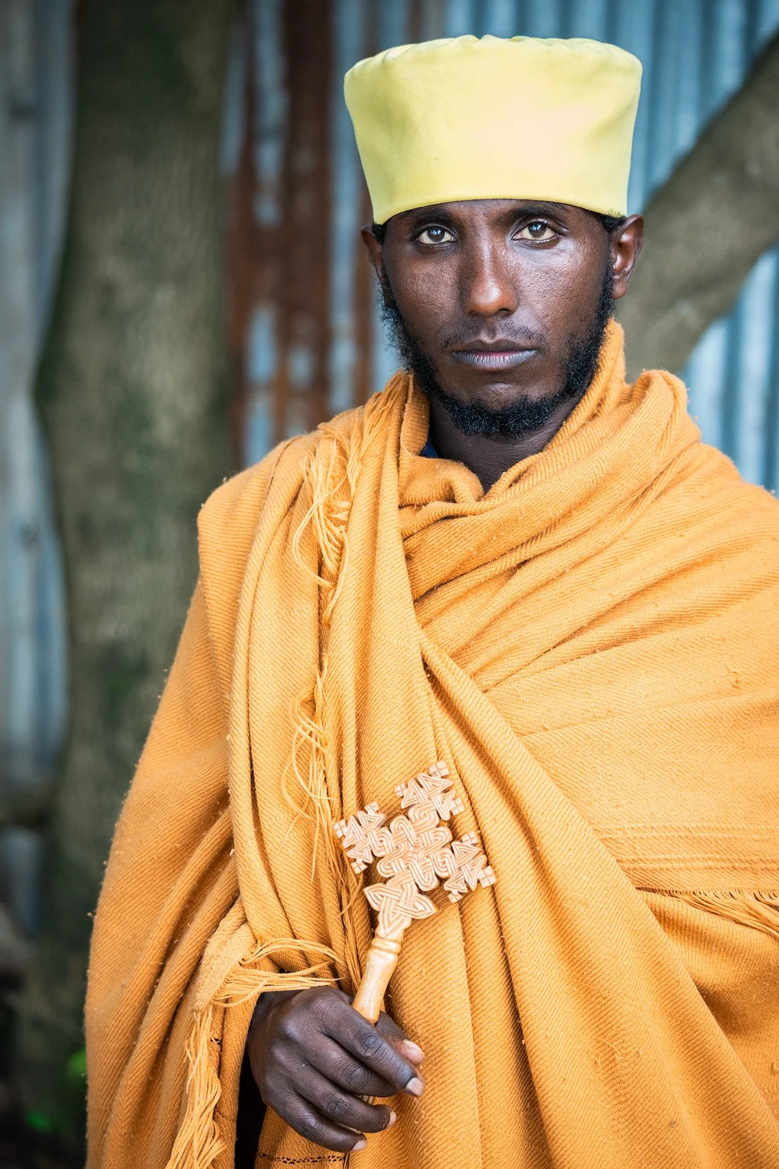 A man dressed in traditional Maasai attire, wearing a yellow headpiece and a yellow shuka, holding a wooden staff with intricate carvings, standing outdoors near a tree.