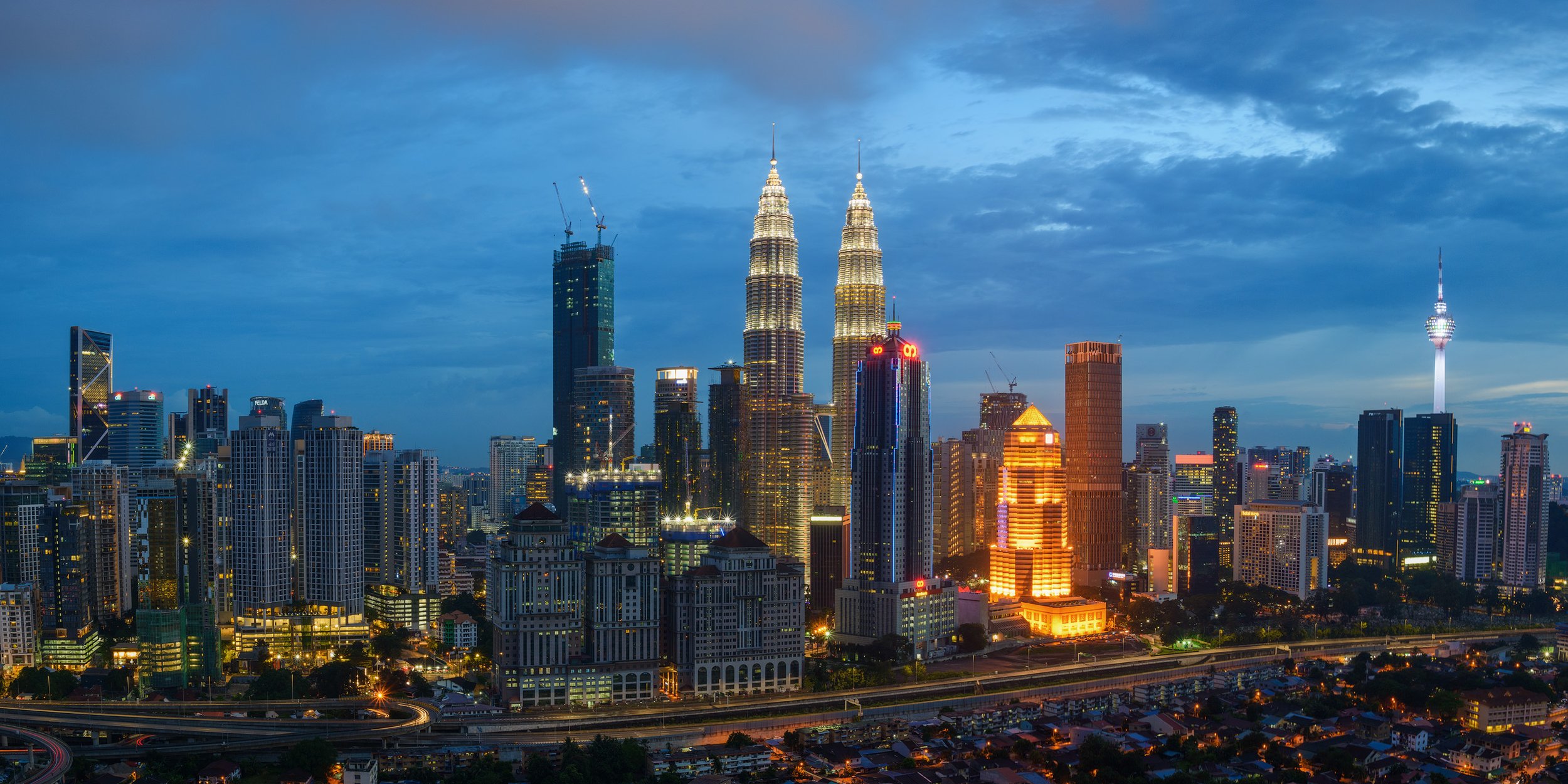 A cityscape of Kuala Lumpur at dusk with illuminated skyscrapers, including the Petronas Twin Towers and the Kuala Lumpur Tower, under a partly cloudy sky.