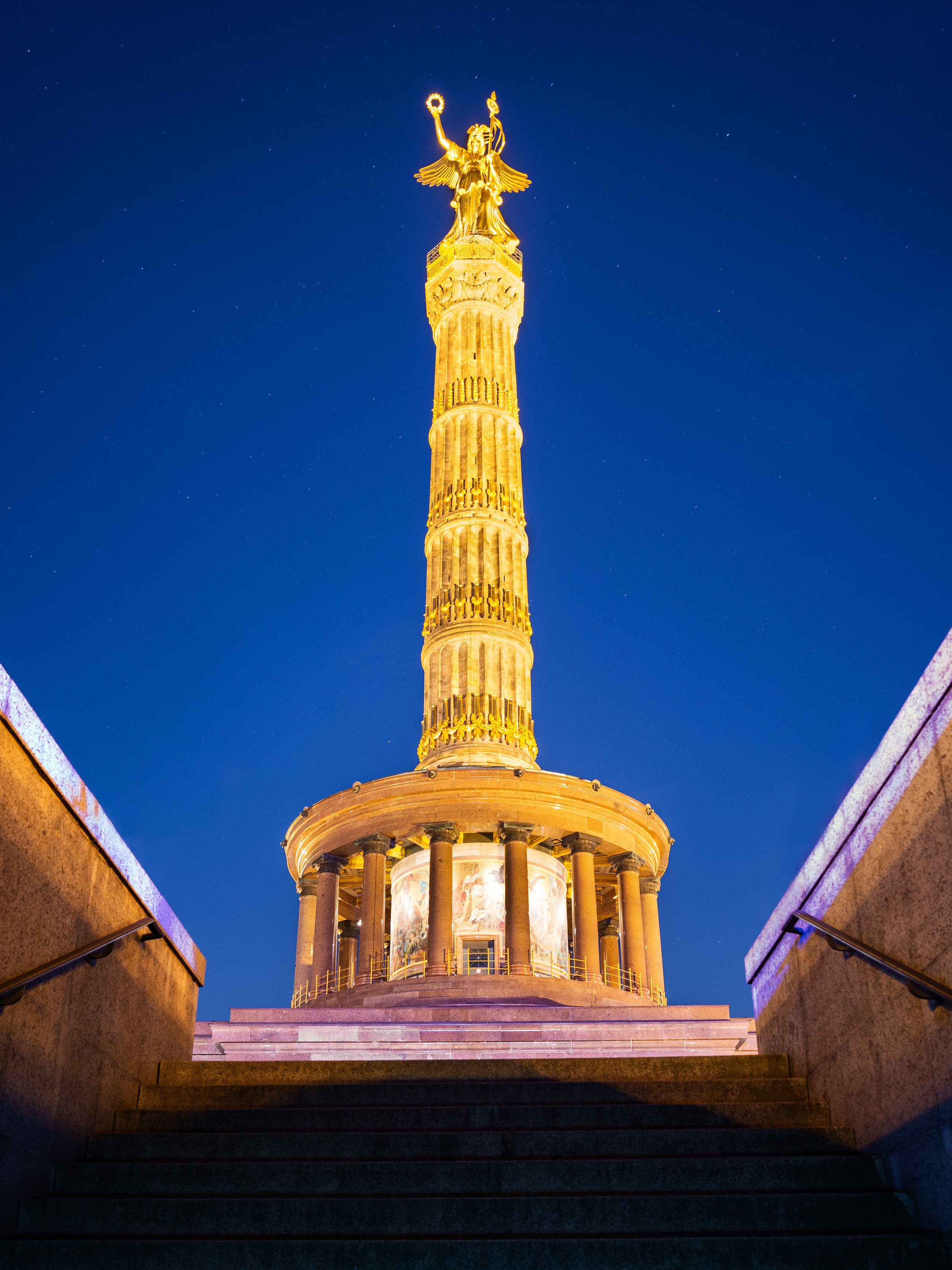 Nighttime view of a tall, golden column topped with a gilded angel statue, with stairs leading up to it and a dark blue sky in the background.