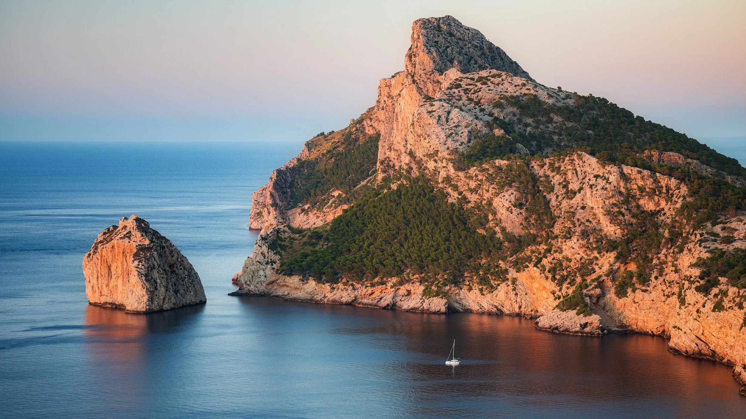 A coastal landscape showing a large rocky hill with green vegetation and a separate large rock formation in the water nearby, with a sailboat in the calm sea at sunset.