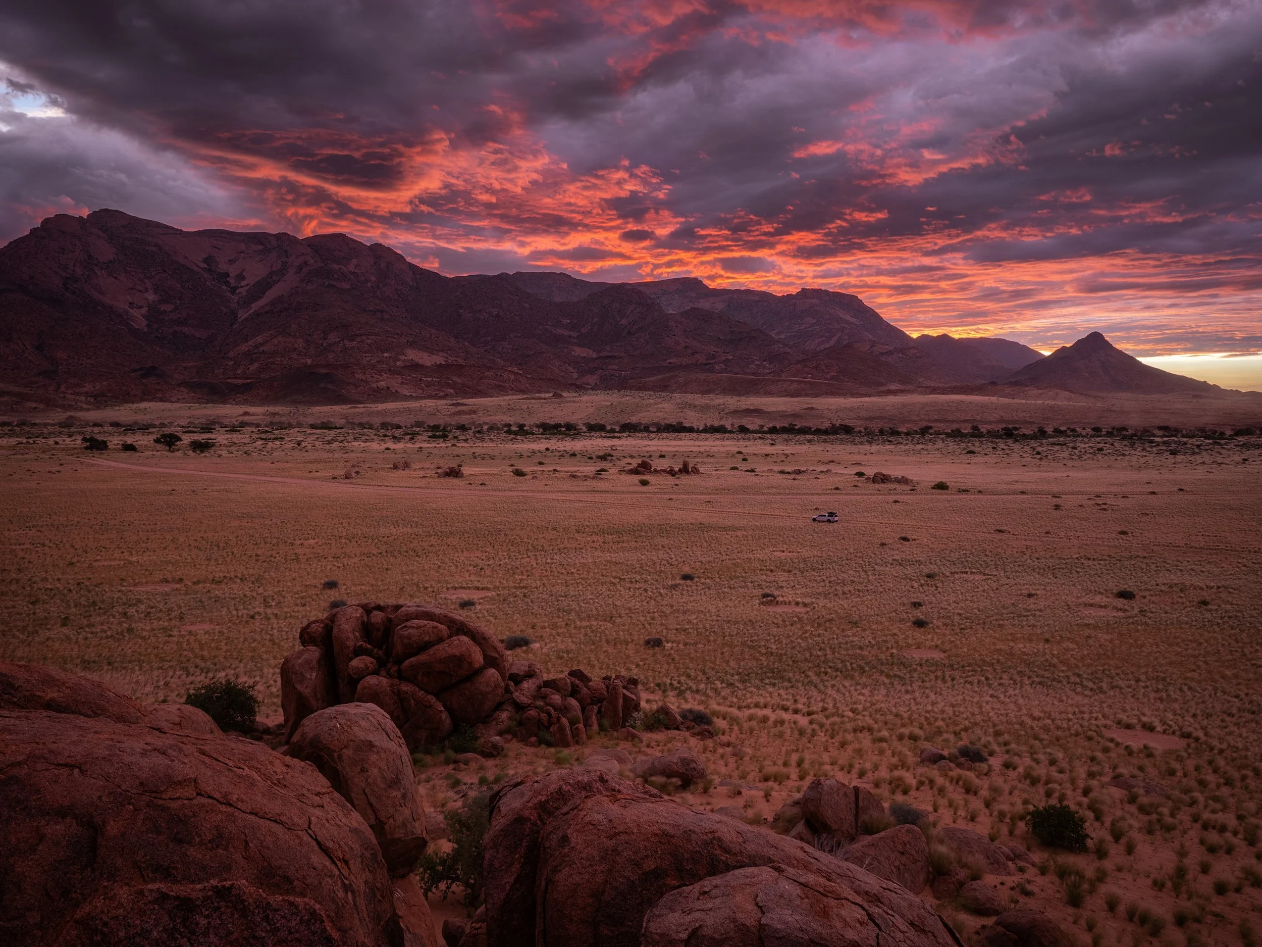 A desert landscape at sunset with rocky hills, sparse vegetation, and a large sky filled with dark clouds tinged with orange and purple hues.