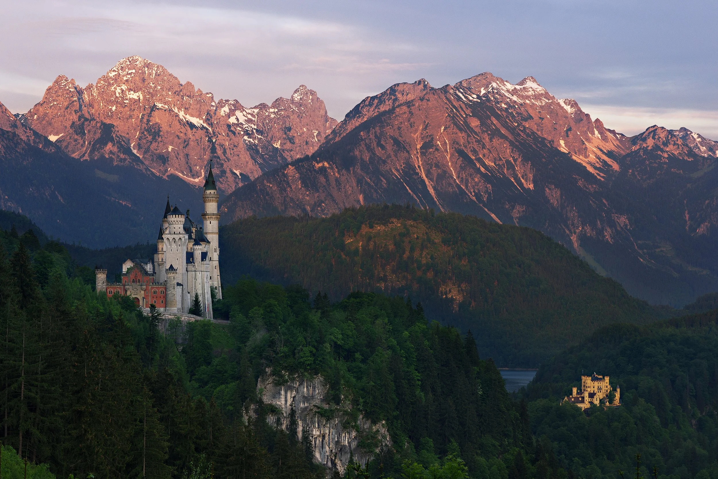 A castle on a hill surrounded by green forests with snow-capped mountains in the background during sunset. Schloss Neuschwanstein. Bayern. Bavaria. Castle. Fairy Tale.