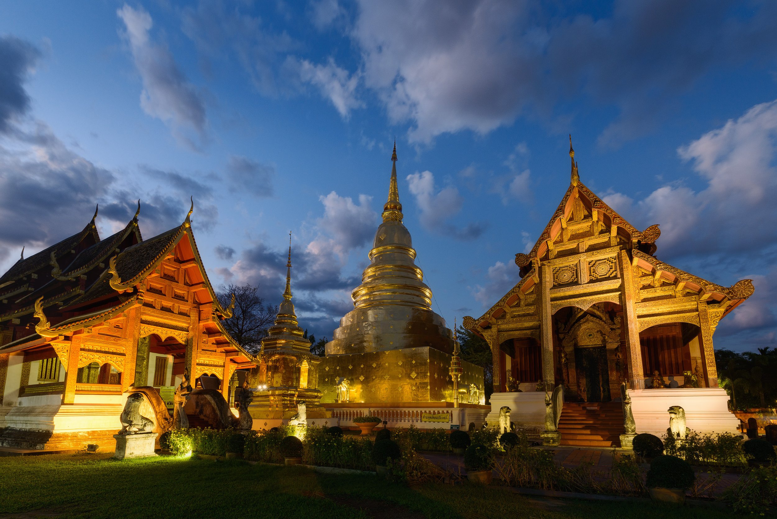 An illuminated traditional Thai temple complex with a golden stupa and ornate wooden buildings against a blue evening sky.
