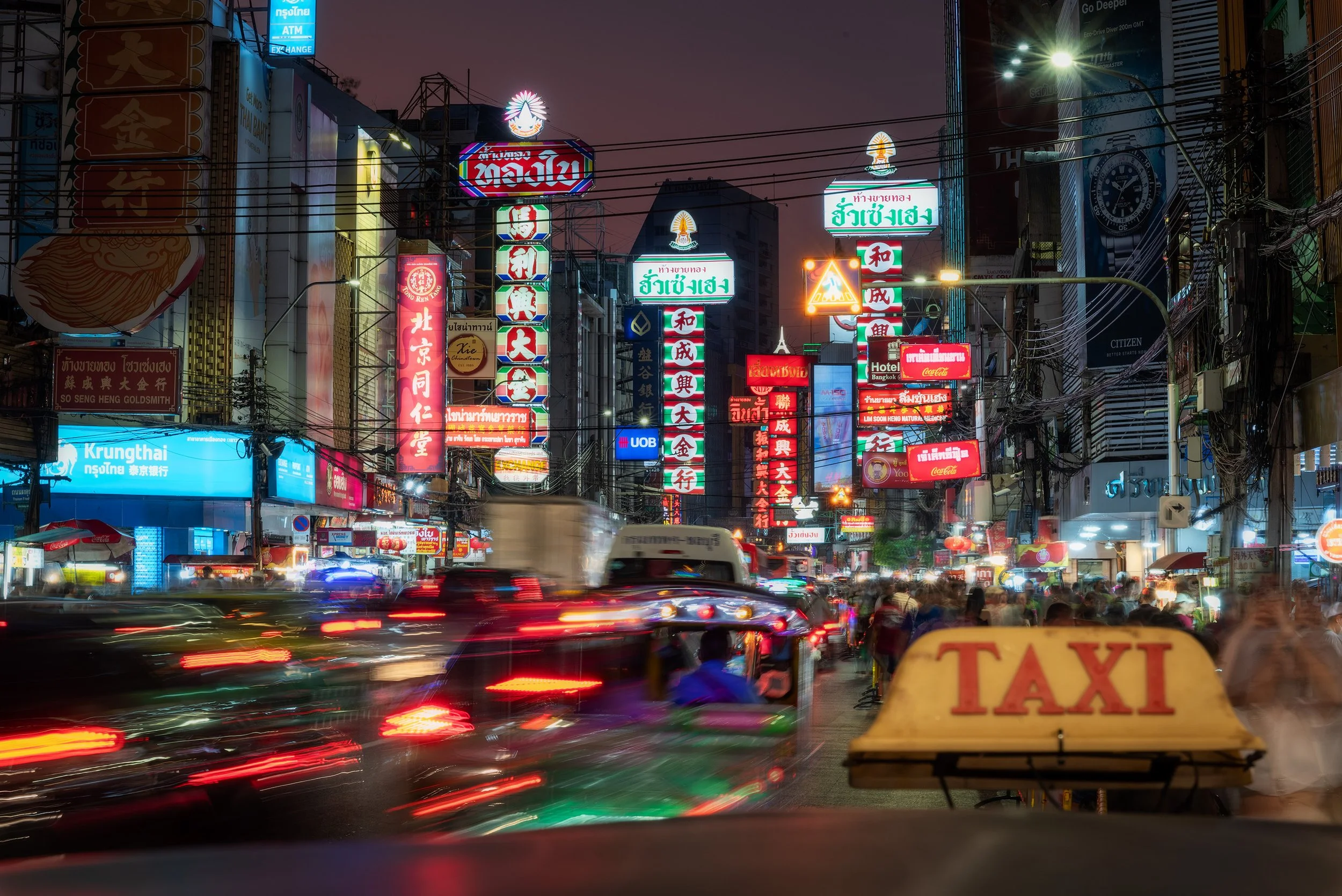 A busy street scene at night in Bangkok, Thailand. With neon signs in various languages including Thai, Chinese, and English, traffic moving quickly with light trails, and a yellow taxi sign in the foreground.