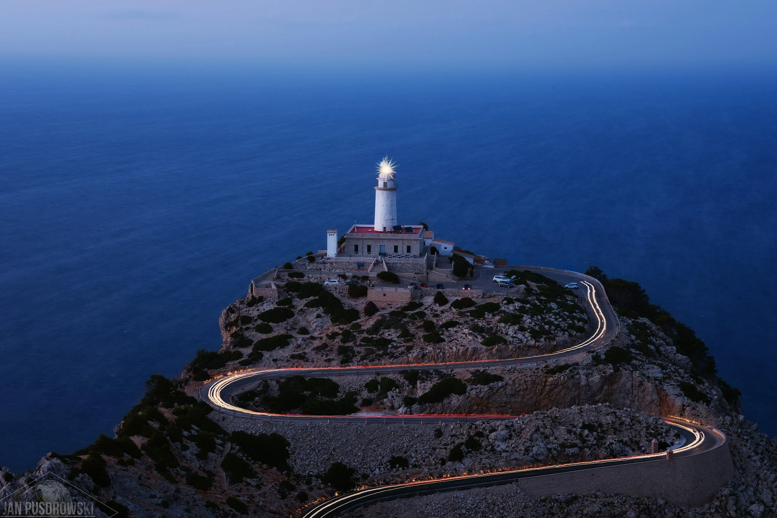 Long exposure photo of a lighthouse on a rocky island with a winding road, cars with light trails, and the ocean in the background during twilight.