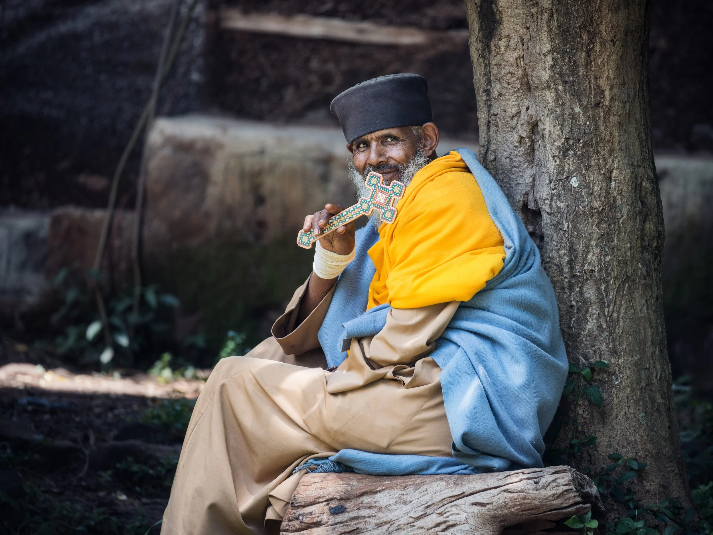 An elderly Ethiopian monk with a gray beard, wearing a black hat, a yellow shawl, and tan clothing, sitting on a log and leaning against a tree, holding a decorated cross near his face and smiling.