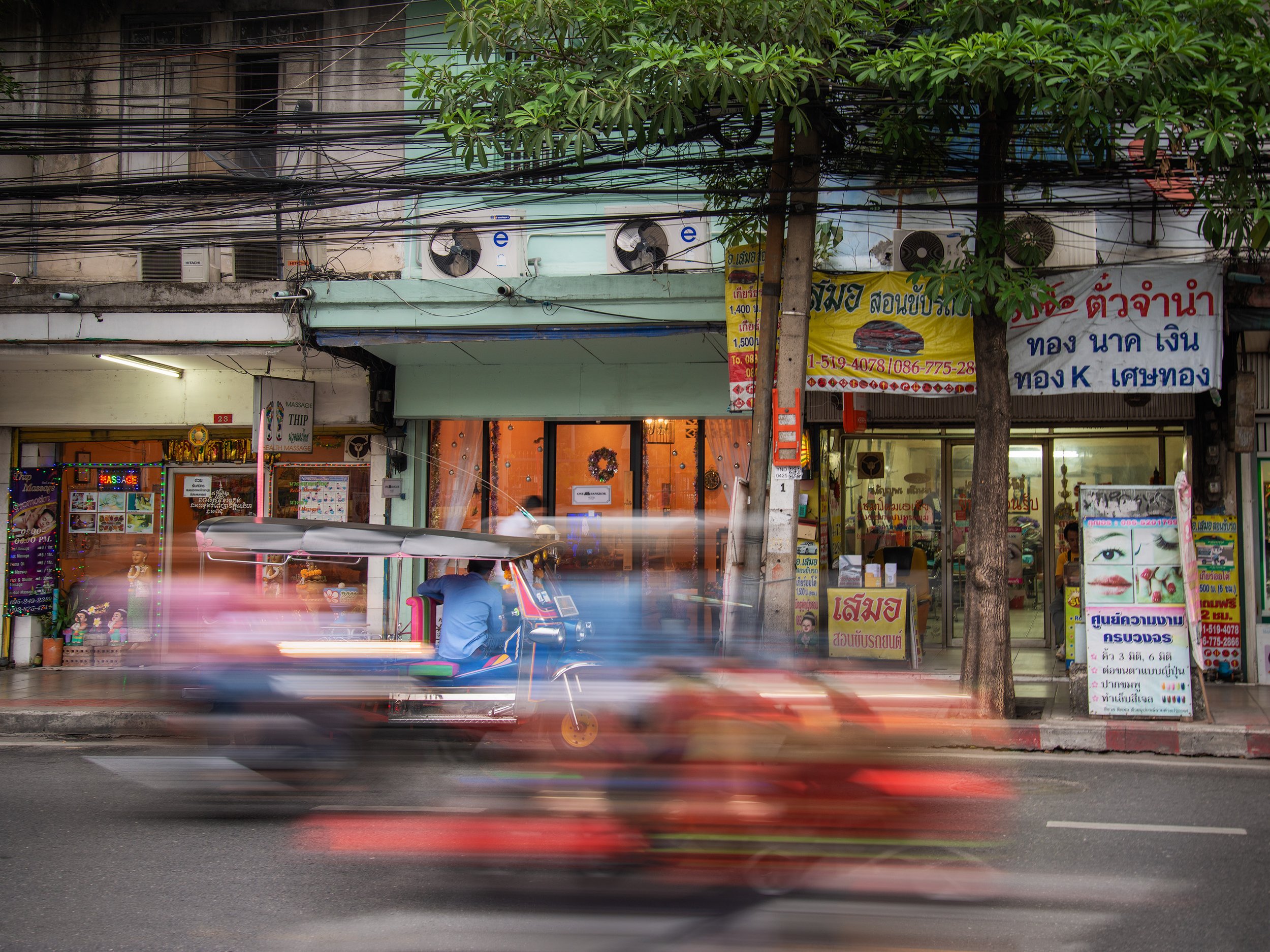 Street scene in an urban area with shops and signs in Thai, a tree, and moving vehicles including a tuk-tuk and a motorcycle.