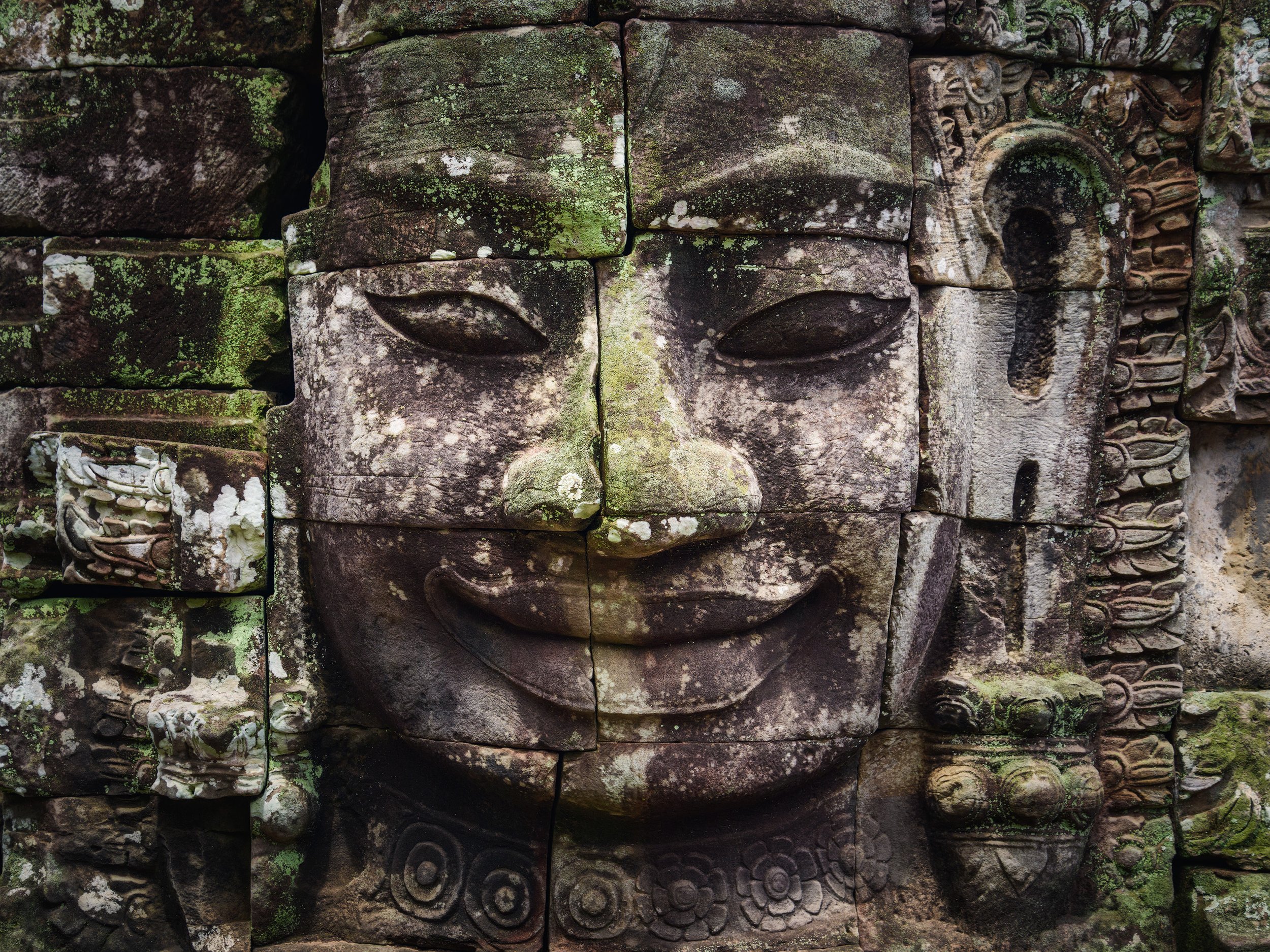 Ancient stone carving of a smiling face with closed eyes, moss-covered and surrounded by detailed carvings.