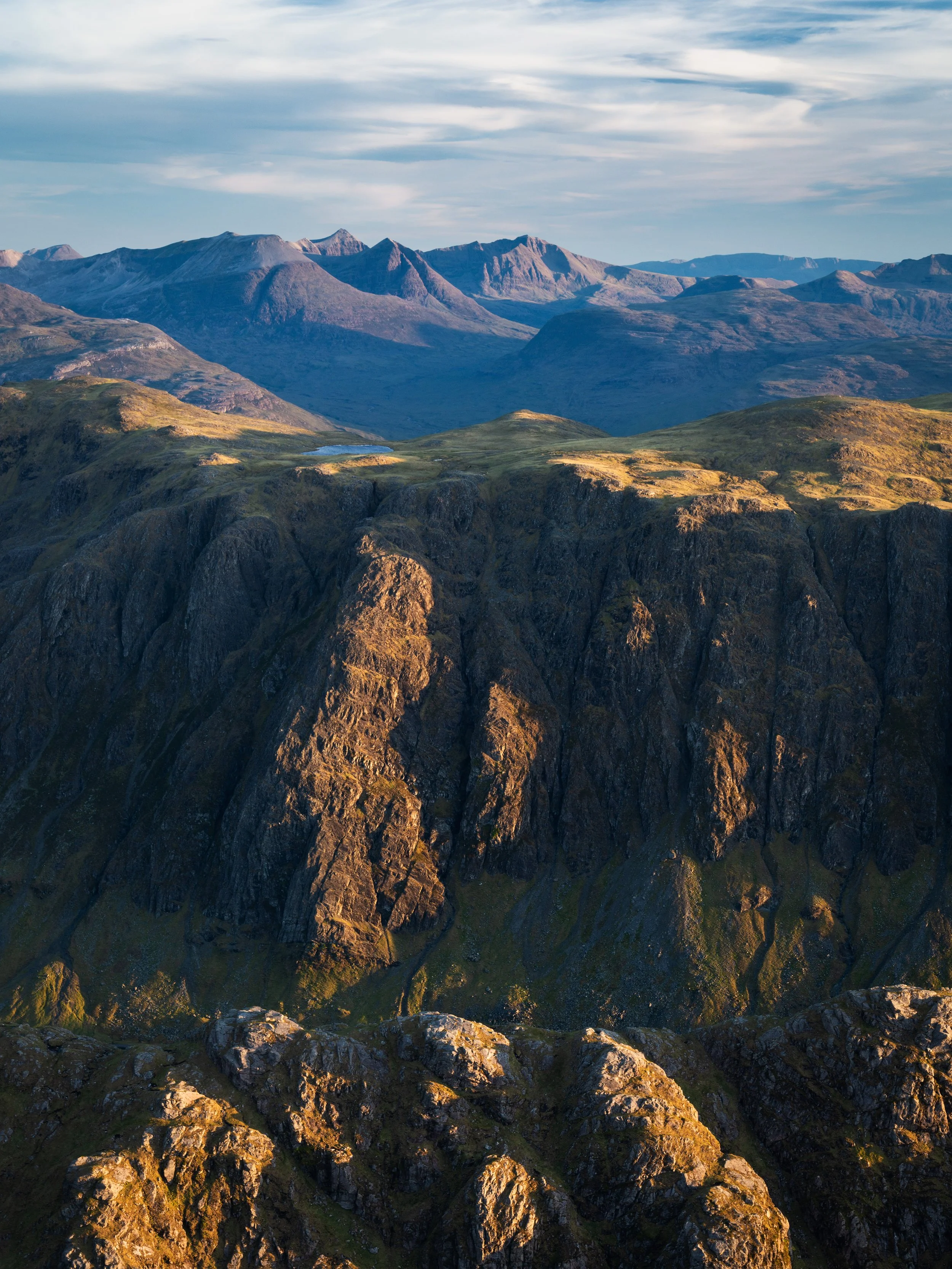 A landscape of rugged mountains with steep cliffs, rolling green hills, and distant mountain peaks under a partly cloudy sky.
