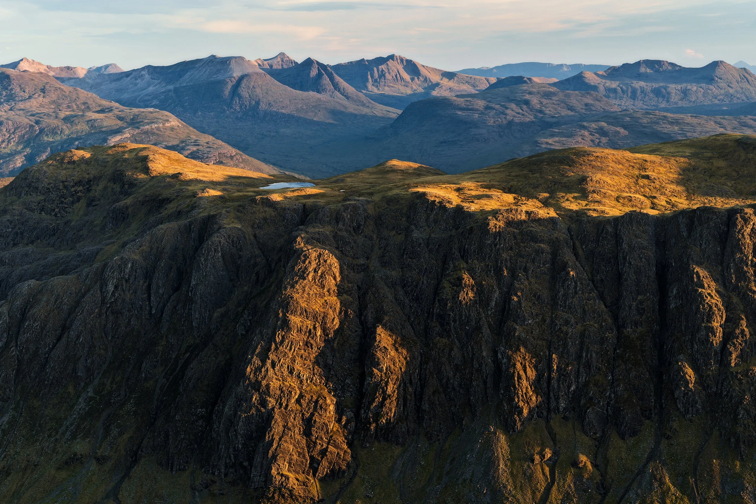 A vast mountain landscape during sunset with rugged dark cliffs in the foreground and rolling green hills, small lakes, and distant peaks in the background.