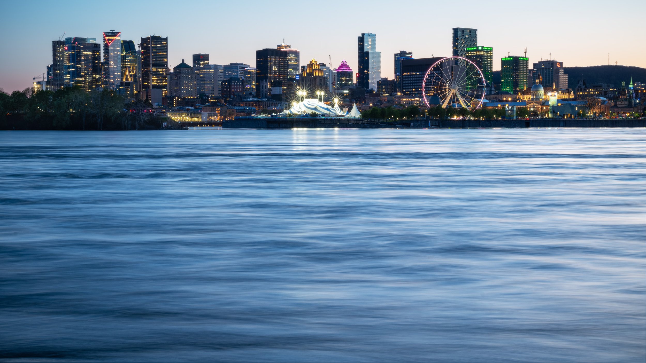 Montreal skyline at dusk with illuminated buildings, a ferris wheel, and a river in the foreground.