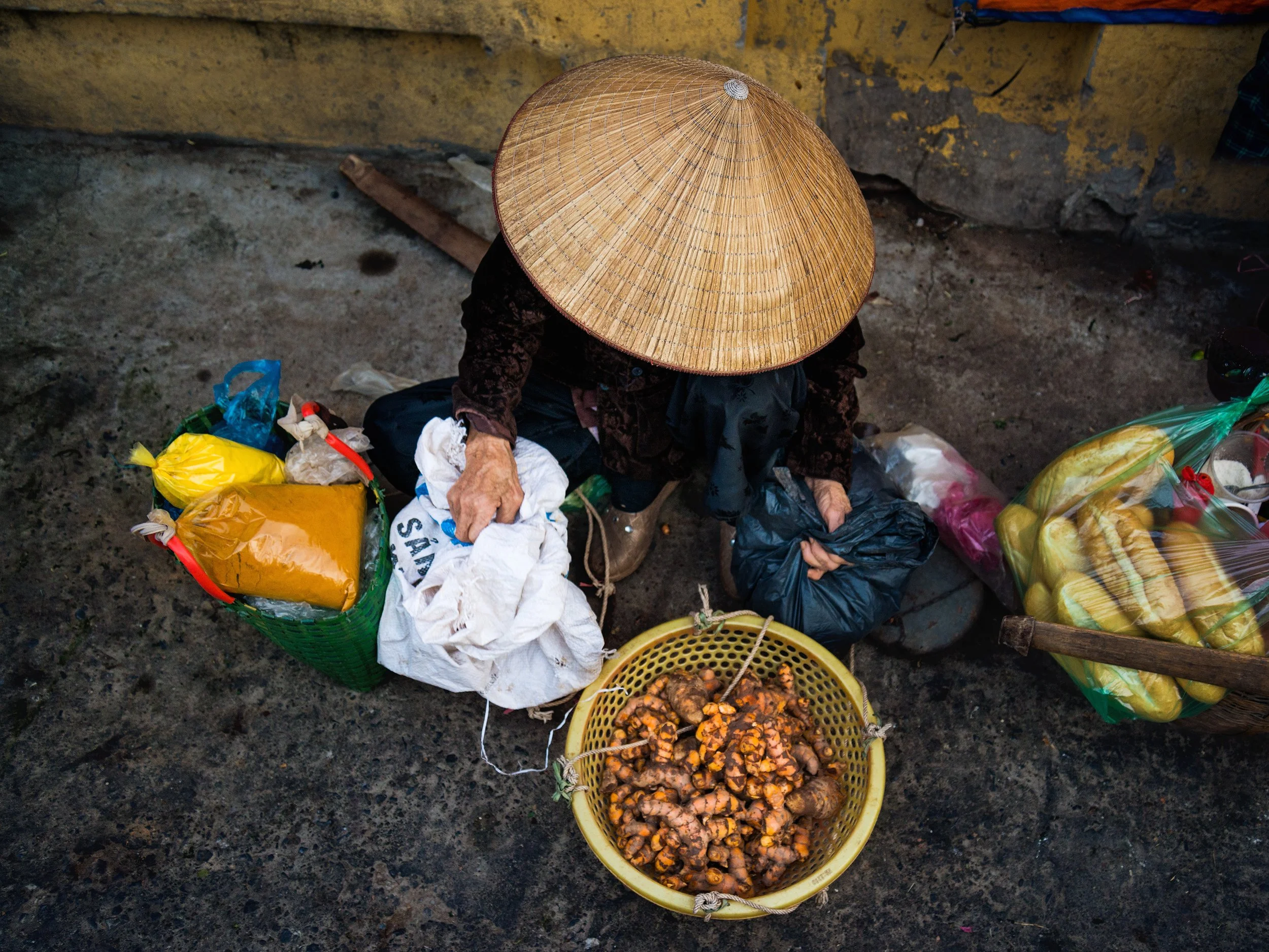 A person wearing a traditional Asian conical straw hat shopping at a street market, surrounded by bags of produce including turmeric, onions, and other vegetables.