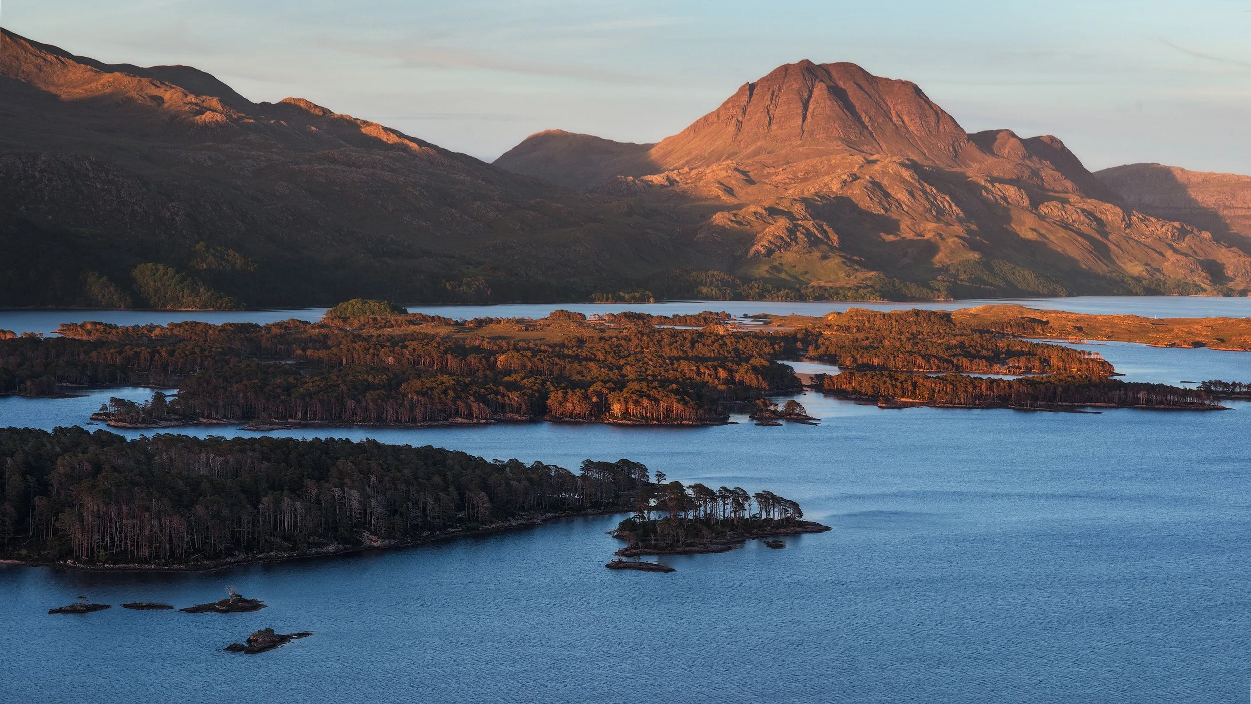 Loch Maree and its small islands, surrounded by forested land, mountains in the background, and a sky with light clouds.