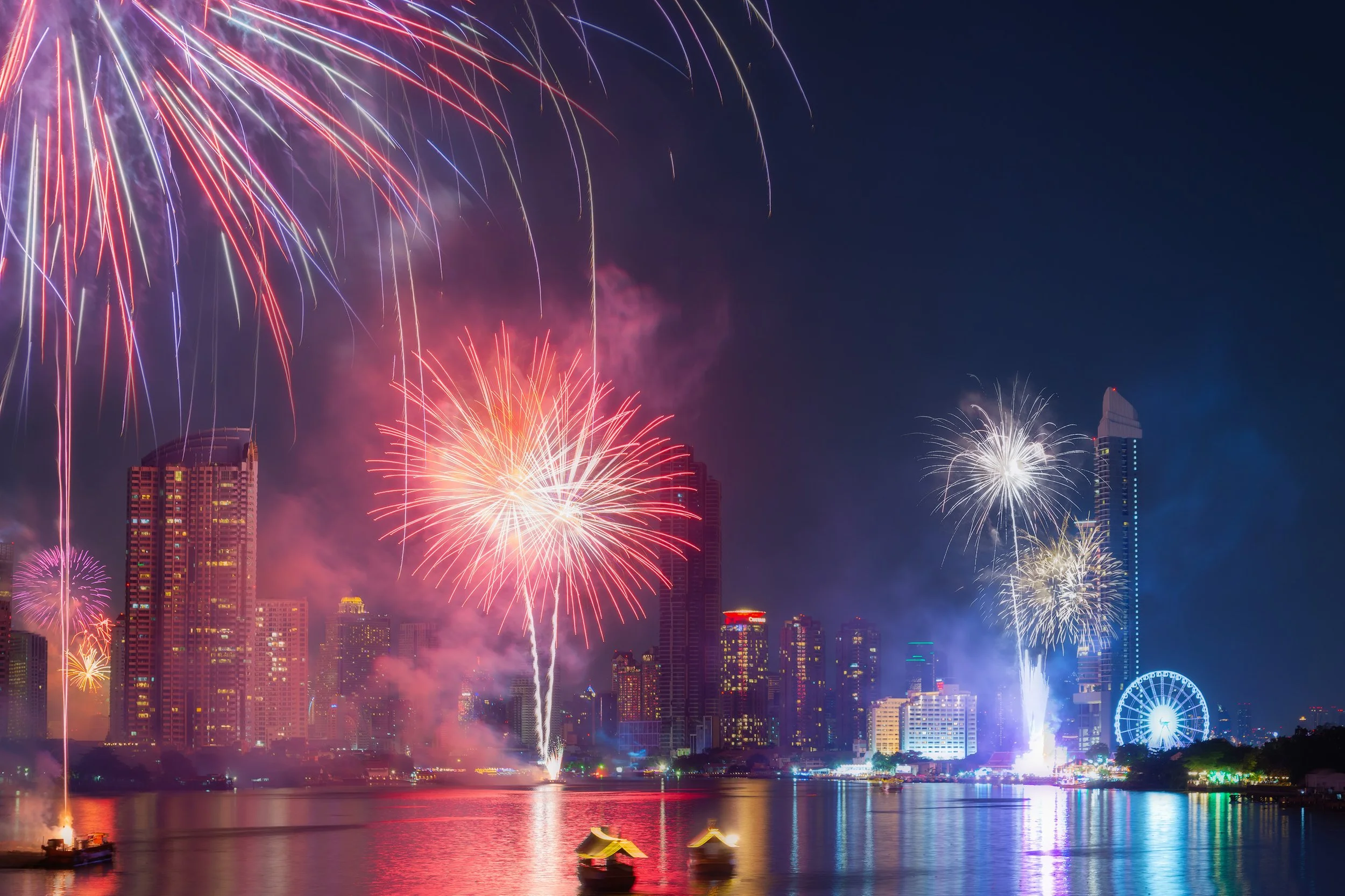 Nighttime cityscape of Bangkok with fireworks over a river, colorful reflections on the water, and a Ferris wheel on the skyline. New Years
