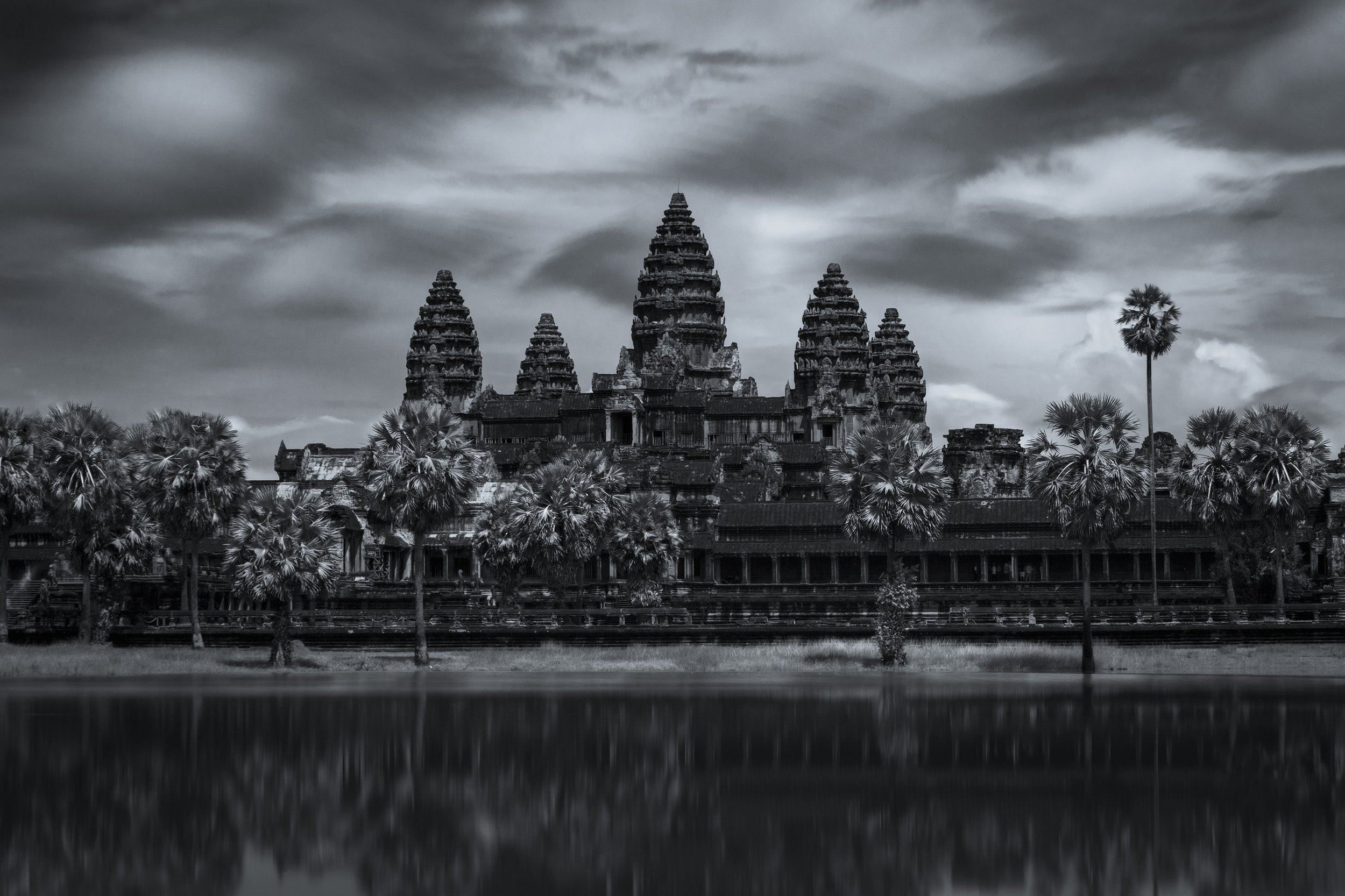 Black and white photo of Angkor Wat temple complex with palm trees and a cloudy sky in the background.