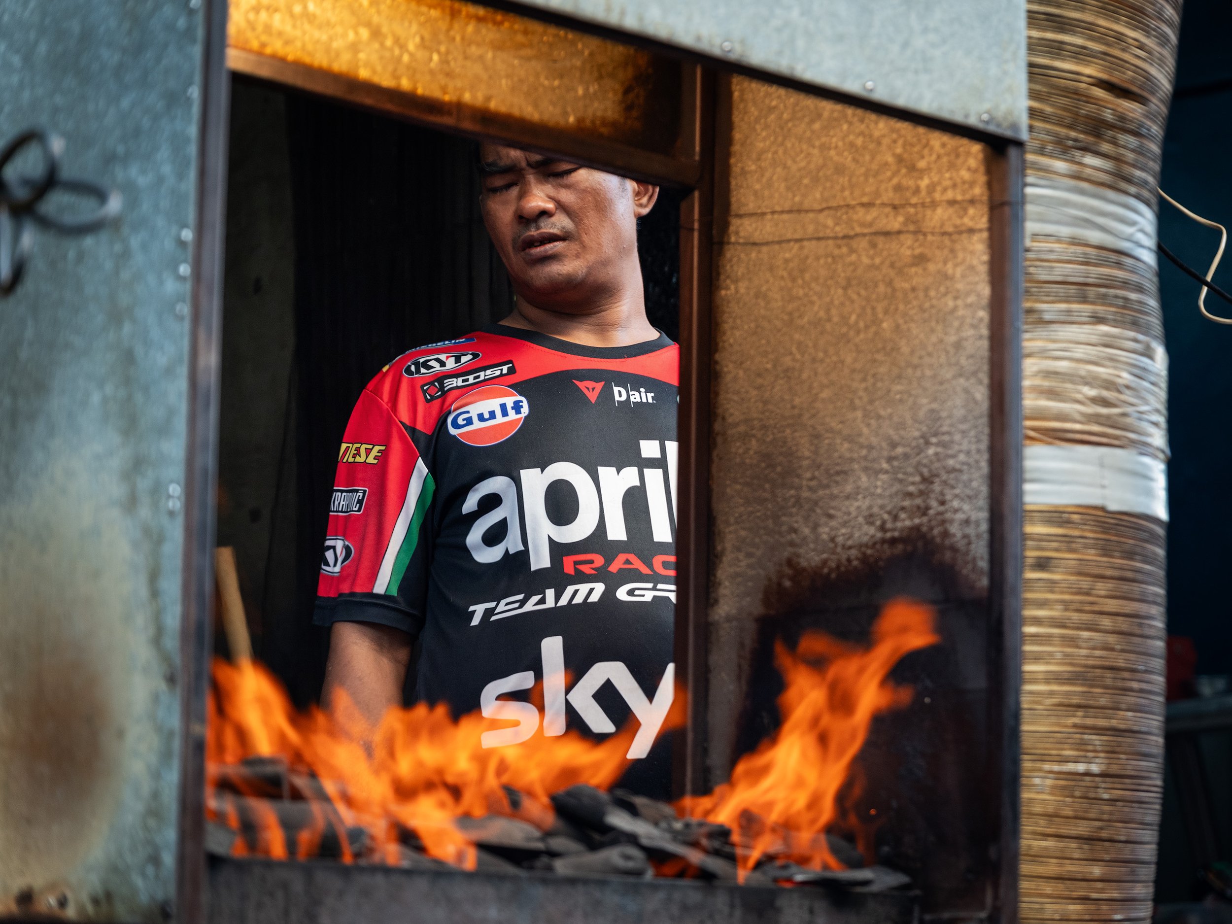 A man wearing a red and black Aprilia racing shirt stands behind a glass window with a fire burning in front of him.