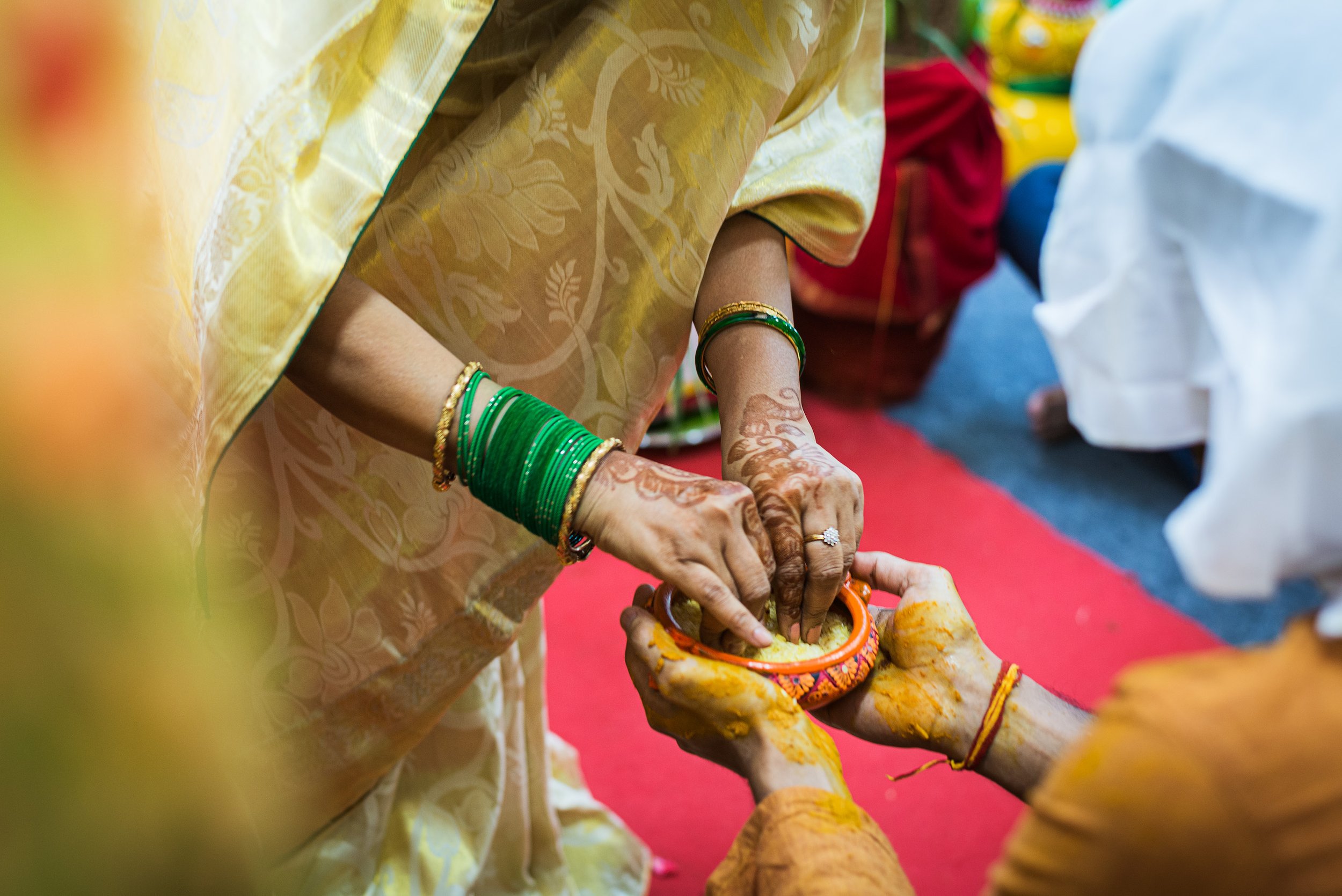 A woman in traditional Indian attire with green bangles and henna on her hands is participating in a ritual involving an orange container with yellow substance, assisted by another person. The setting appears to be a festive or cultural ceremony.