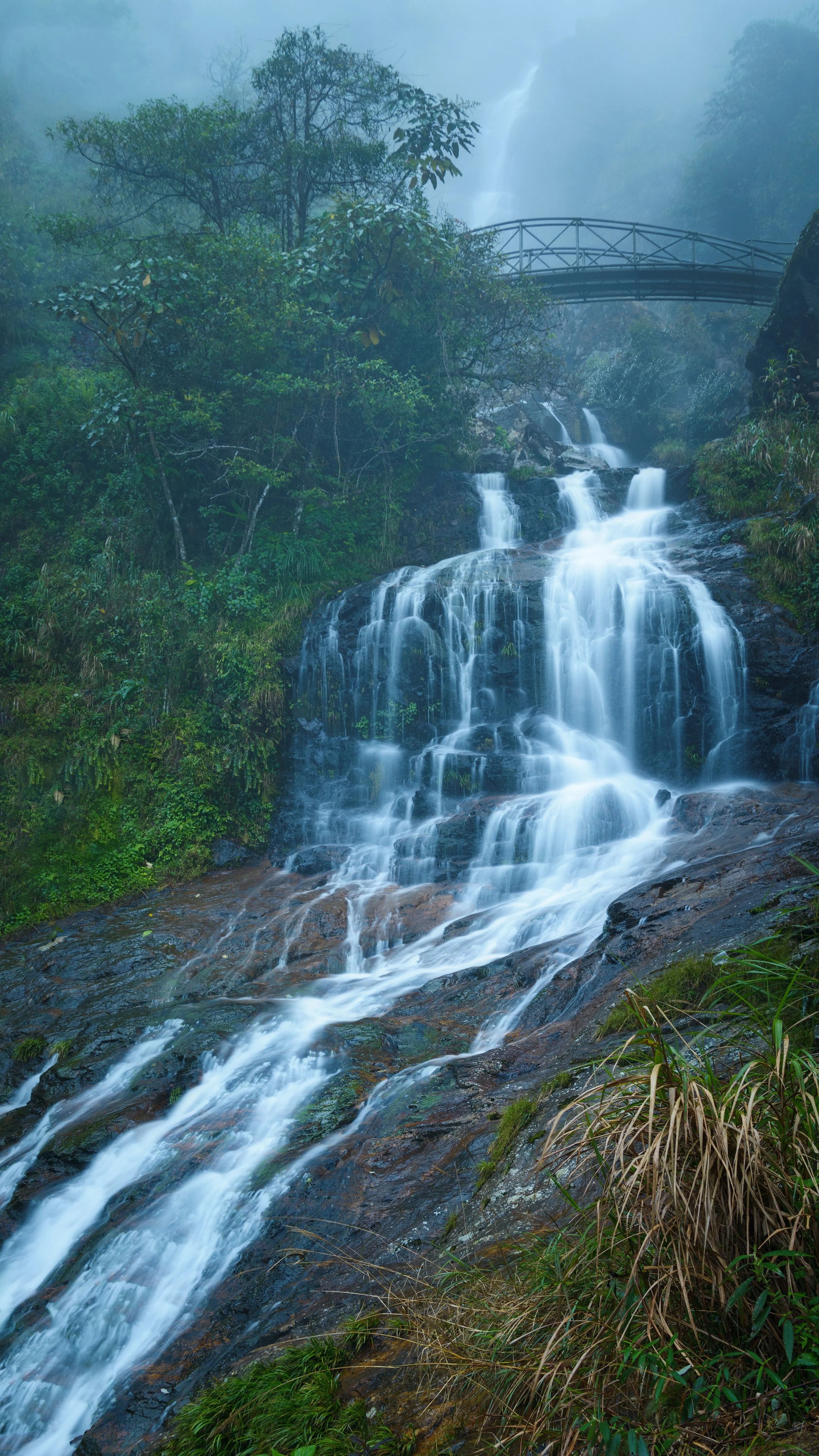 A scenic view of a multi-tiered waterfall flowing over rocks surrounded by lush green vegetation with a misty atmosphere and a bridge at the top in the background. Vietnam, Viet Nam, Sapa.