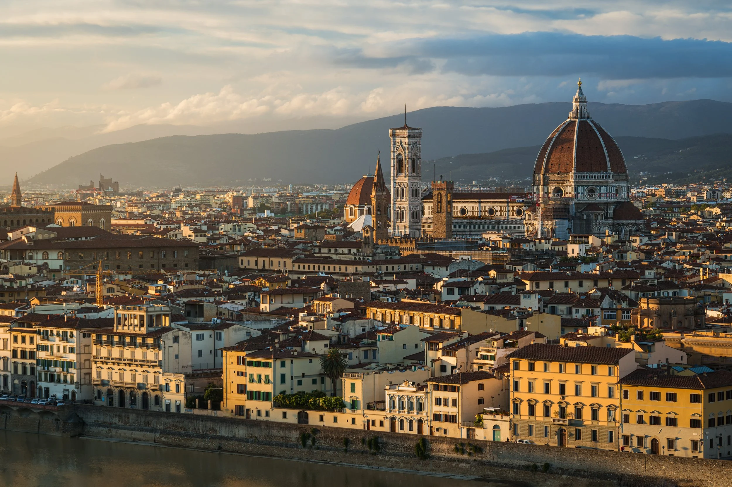 A cityscape of Florence, Italy, during sunset featuring the Florence Cathedral with its large dome, the bell tower, and numerous historic buildings with red-tiled roofs, set against a backdrop of rolling hills and mountains.