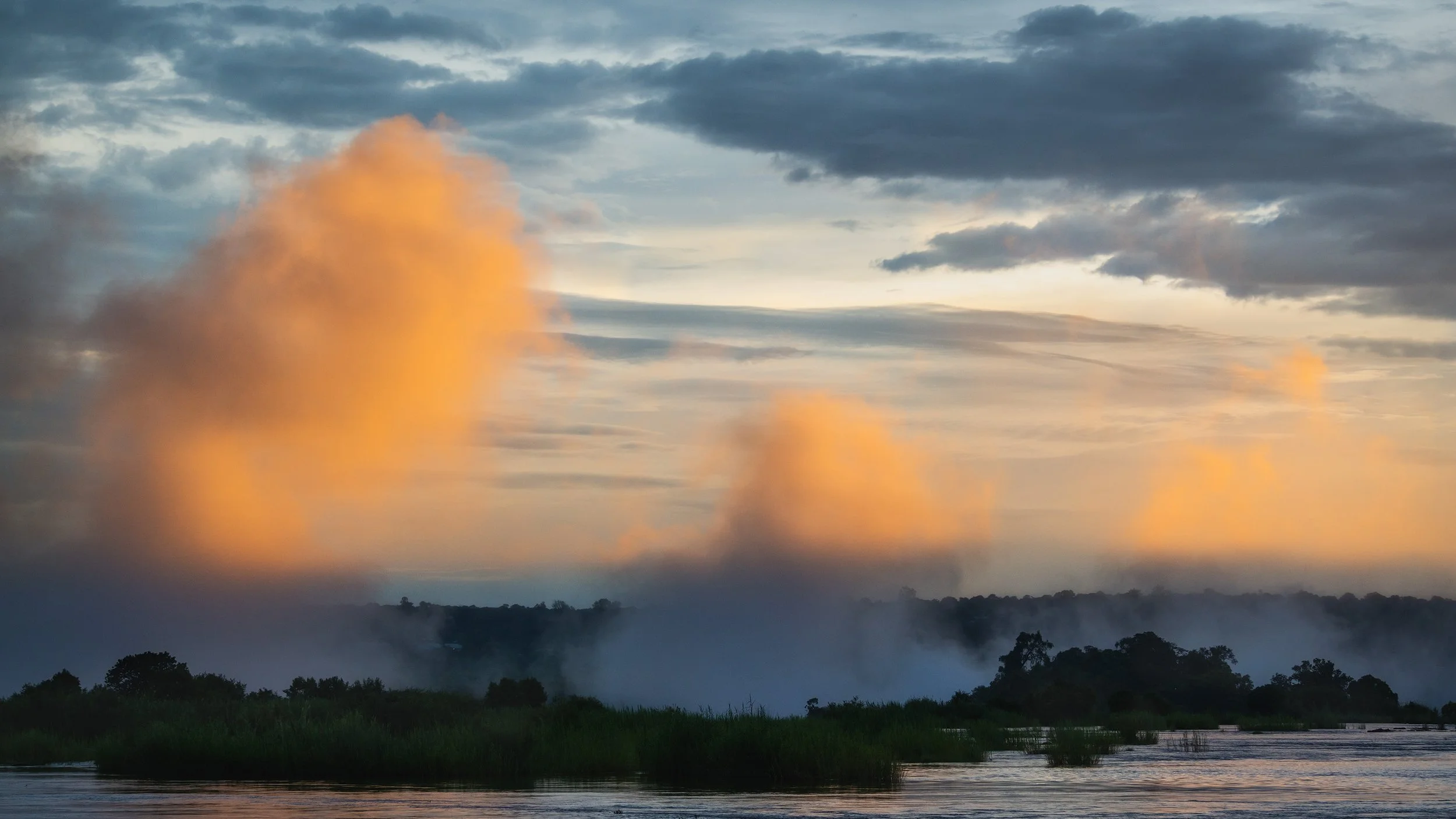 Landscape scene with a river, green vegetation, trees in the distance, and a sky with dark clouds and orange-tinted clouds near the horizon.