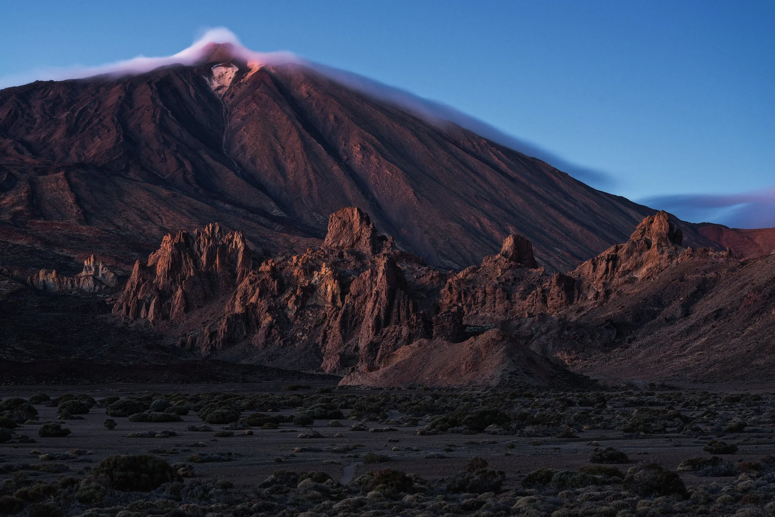 A mountain with a volcanic cone partially covered by clouds, with rocky formations and sparse desert vegetation in the foreground.