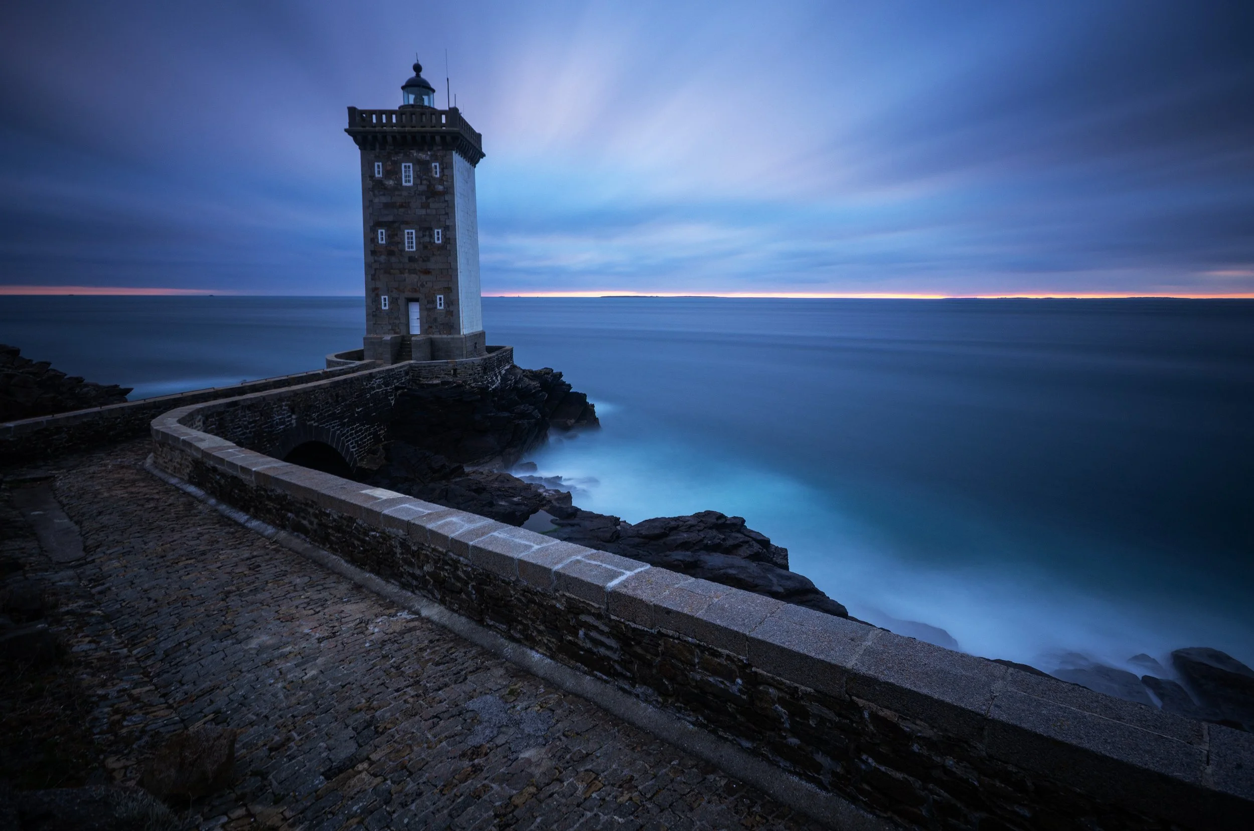 A lighthouse on a rocky coastline at dusk, with a cloudy sky and calm ocean waves.