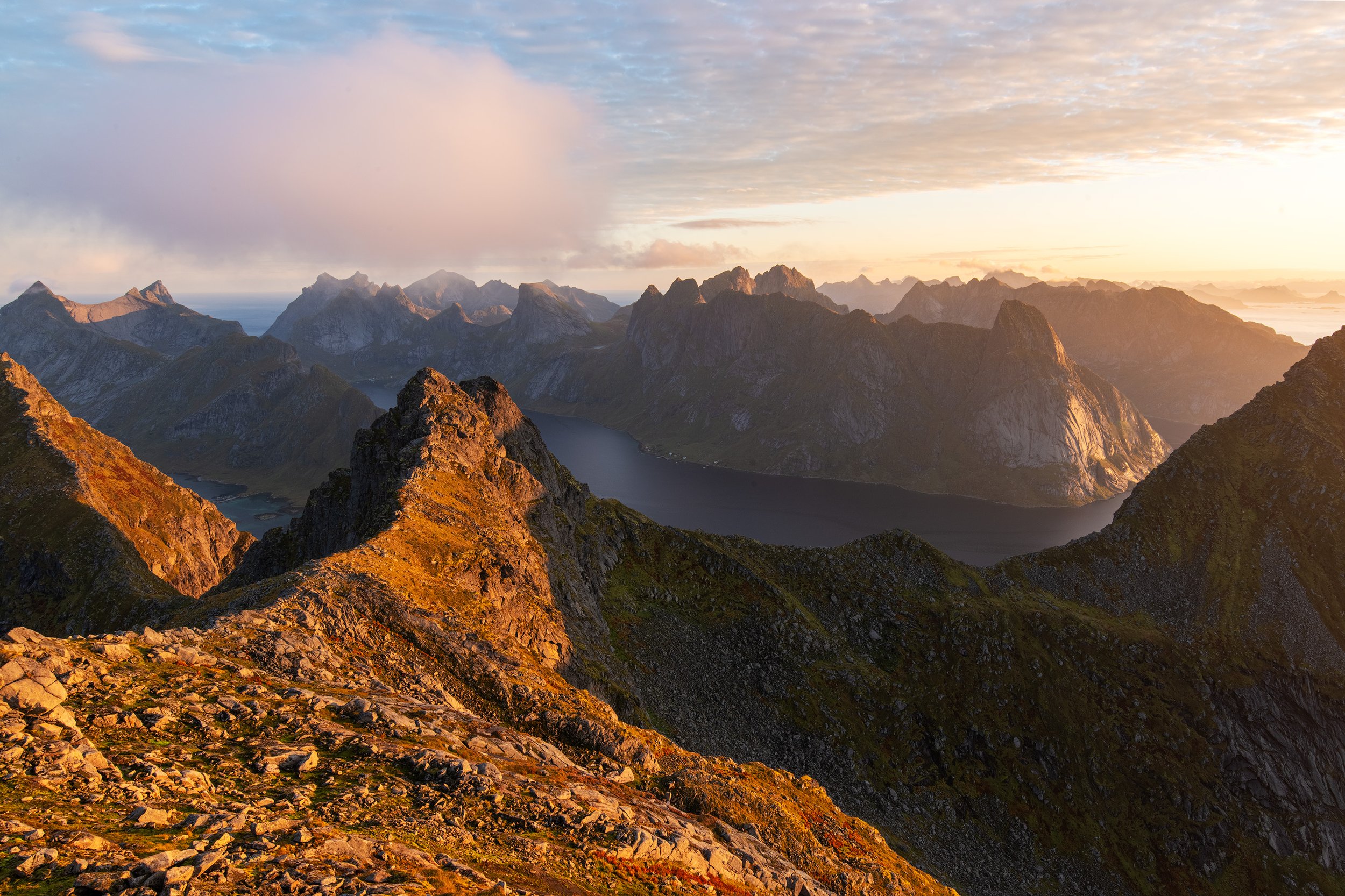Sunrise over a mountain range with rugged peaks, a lake in the valley, and a partly cloudy sky. Lofoten, Norway.