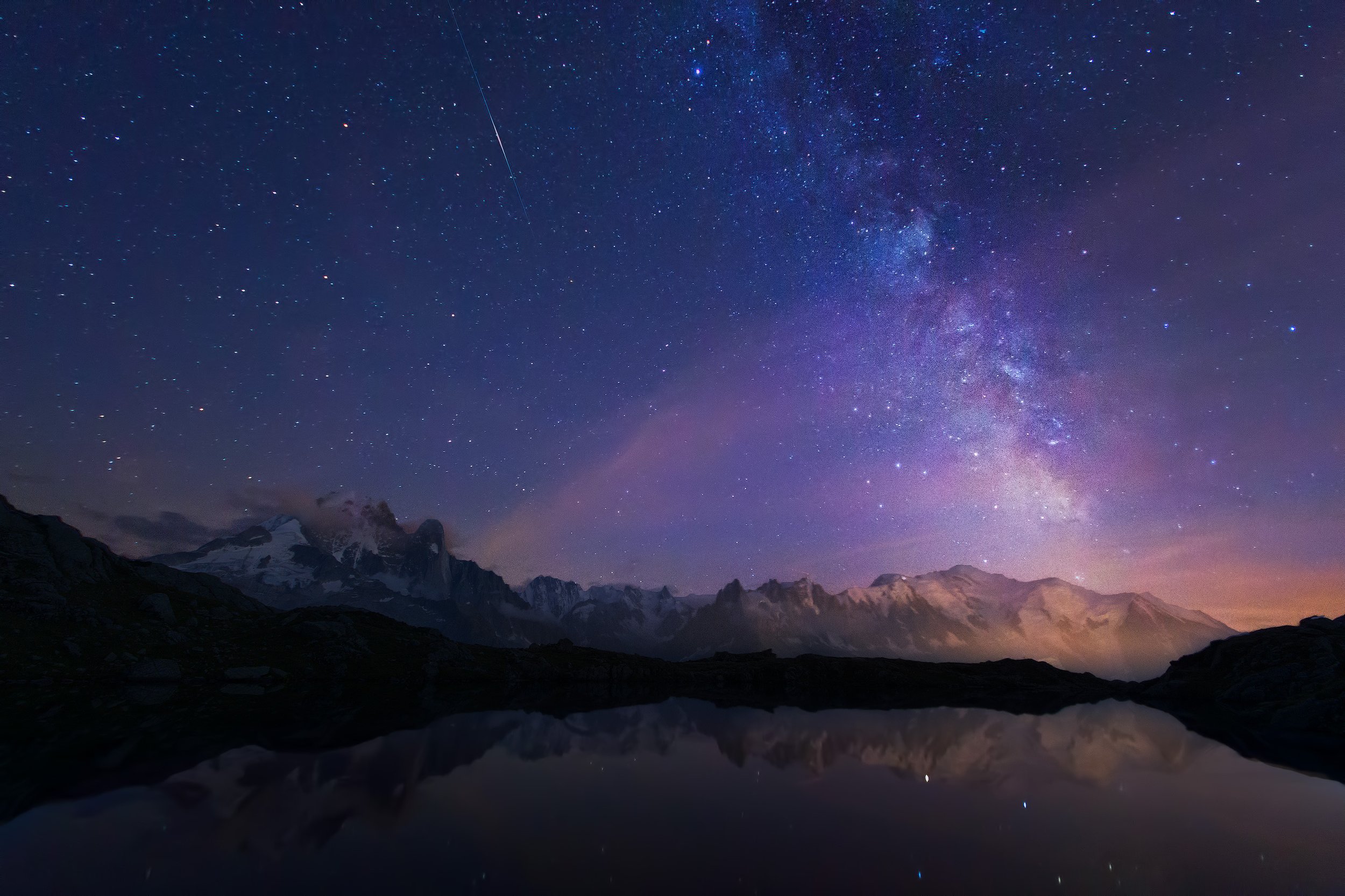 Night sky filled with stars and the Milky Way galaxy over mountains and a reflective body of water.