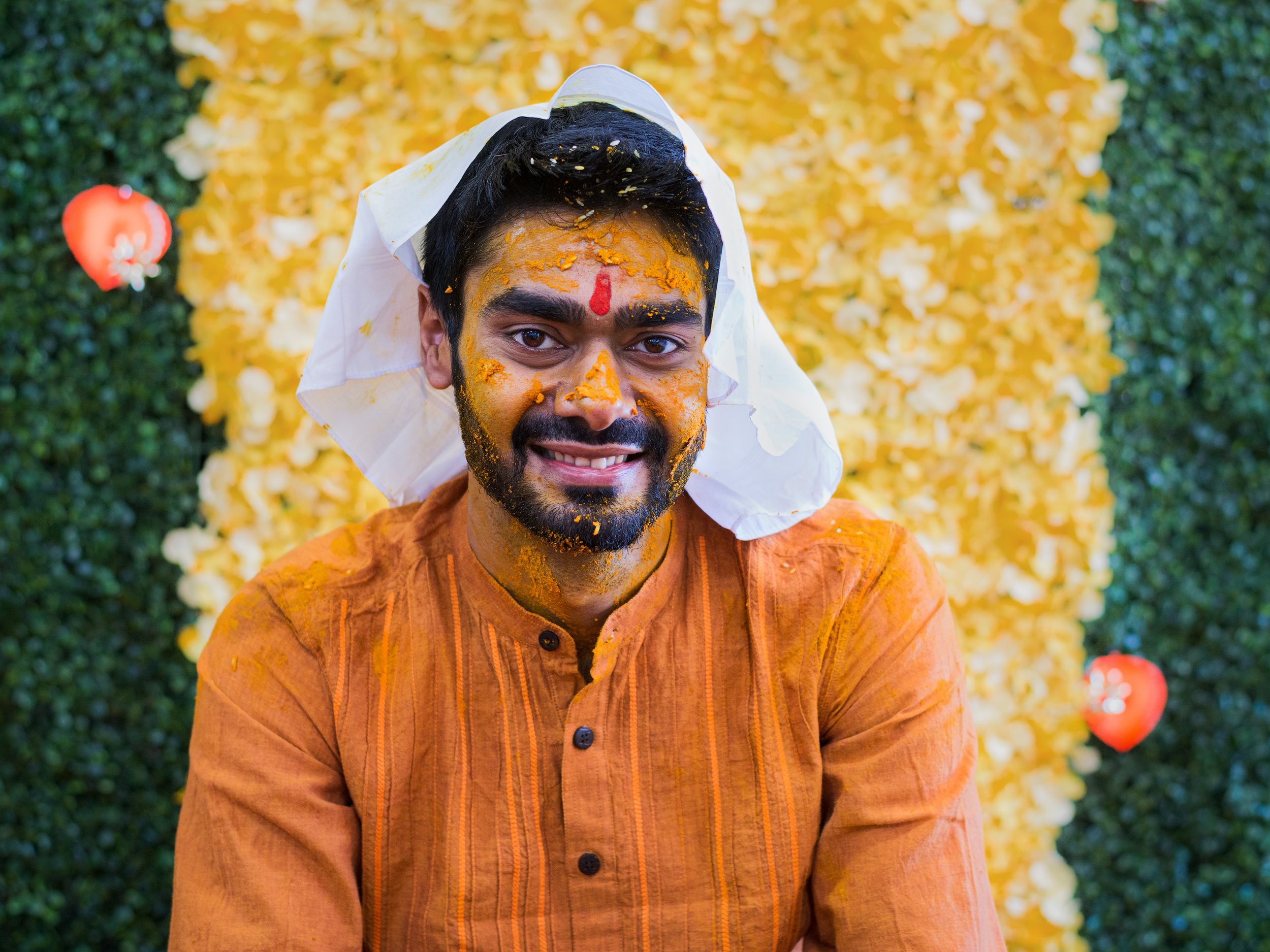 A man with yellow and orange powder on his face and clothing, smiling, with a festive backdrop of yellow and green decorations and red balloons.